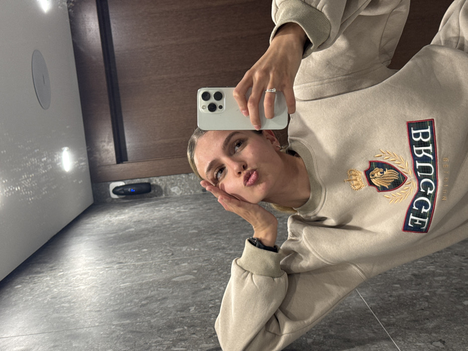 The image is a mirror selfie taken indoors. A female subject, with light brown hair pulled back, is visible from the chest up, reflected in a mirror. She is wearing a beige long-sleeved sweatshirt adorned with an embroidered crest featuring a lion's head and a crown, below which the inverted words "BRUGGE" and "BELGIUM" are displayed. Her right hand holds a light-colored smartphone, aimed towards the mirror, with a ring visible on her right ring finger. Her left hand is positioned with fingers splayed against her left temple. Her lips are pursed. The reflected background consists of a modern interior featuring a dark grey, possibly marble or granite, wall on the left, transitioning to a dark wood-paneled wall on the right. A black, vertical, wall-mounted device with a blue indicator light is affixed to the grey wall. The ceiling is white with visible recessed lighting. The specific location within Tbilisi, Georgia, is not identifiable from the visual evidence.