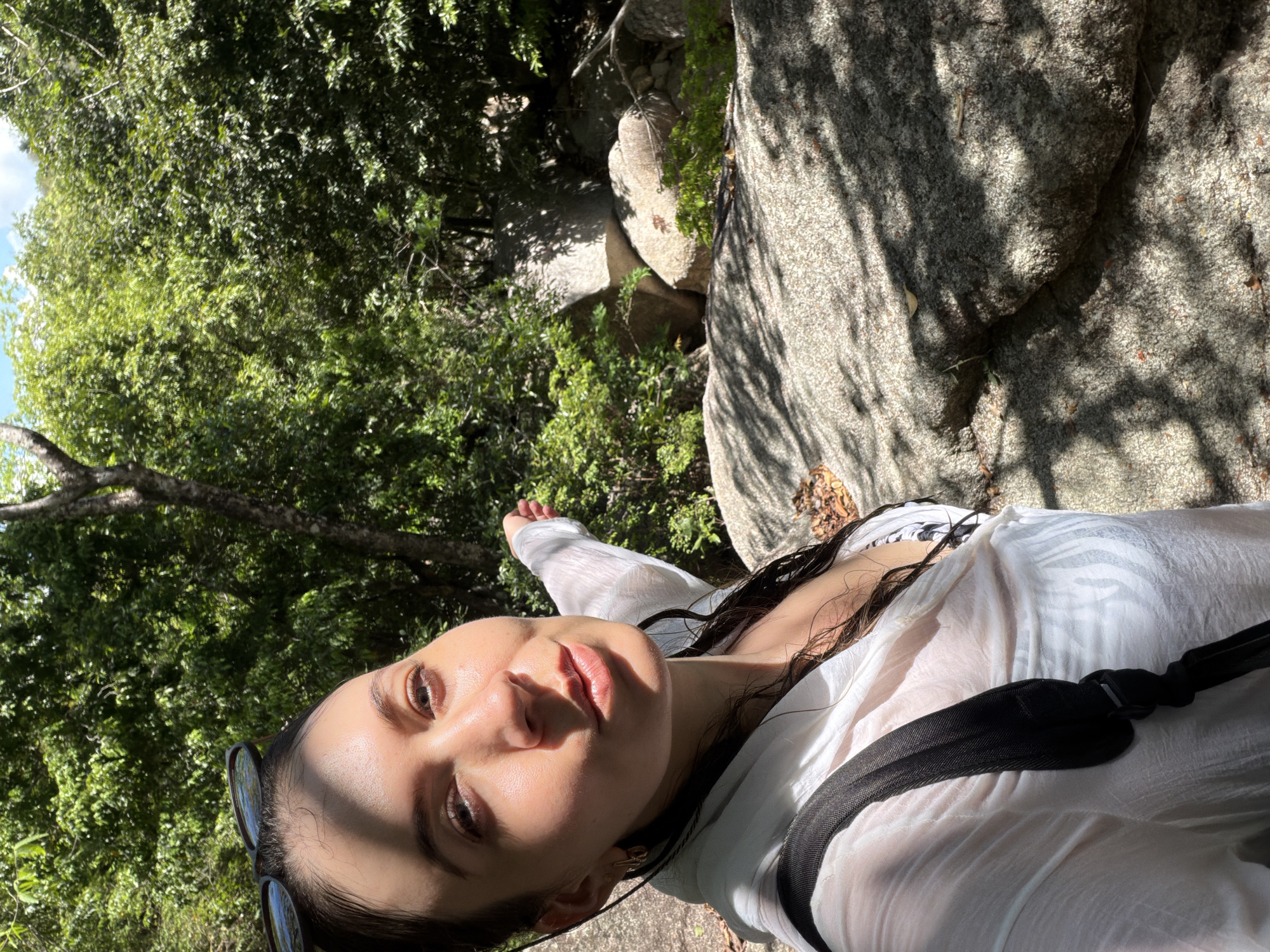 A young woman is taking a selfie in a natural environment. She is wearing a sheer white cover-up over a patterned bikini. Sunglasses are perched on her head. Her hair is wet and dark. She is pointing to her right with her left hand, towards a rocky area with dense green foliage. The lighting suggests a sunny day, with dappled shadows from trees falling on the rocks and the woman. The location is identified as Hoa Thang, Vietnam.
