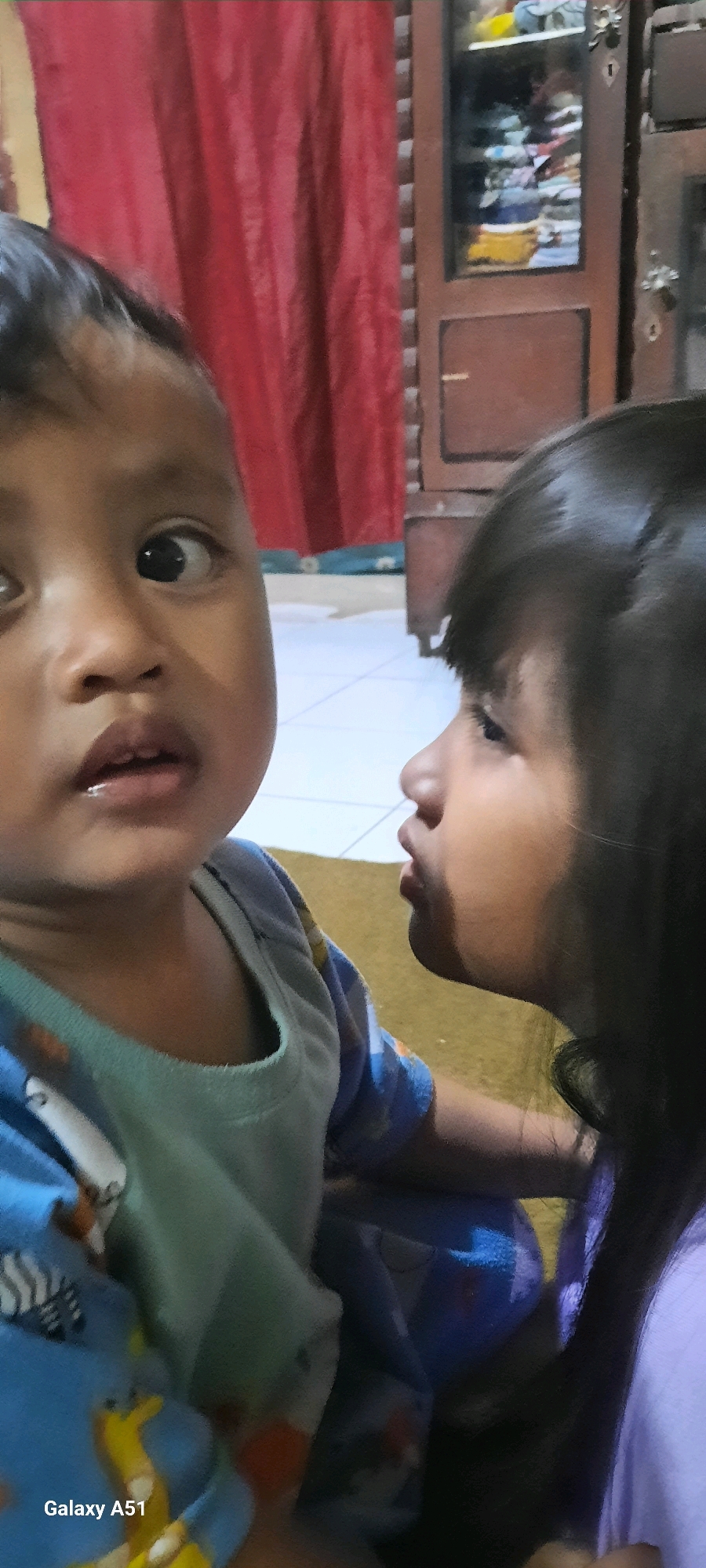 Two young children, a boy and a girl, are positioned closely together in the foreground of the image. The boy, on the left, is looking directly at the camera with wide eyes and his mouth slightly open, revealing his teeth. He is wearing a blue shirt with a light green undershirt, featuring cartoon characters. The girl, on the right, is facing the boy, with her head turned towards him and her mouth pursed as if about to kiss him. Her dark hair is visible. In the background, a red curtain hangs to the left, and a wooden cabinet with glass doors filled with various items is visible on the right. The floor appears to be tiled. The image also has a watermark in the bottom left corner that reads "Galaxy A51". The scene appears to be indoors, likely in a residential setting within the city of Ploso, Indonesia.