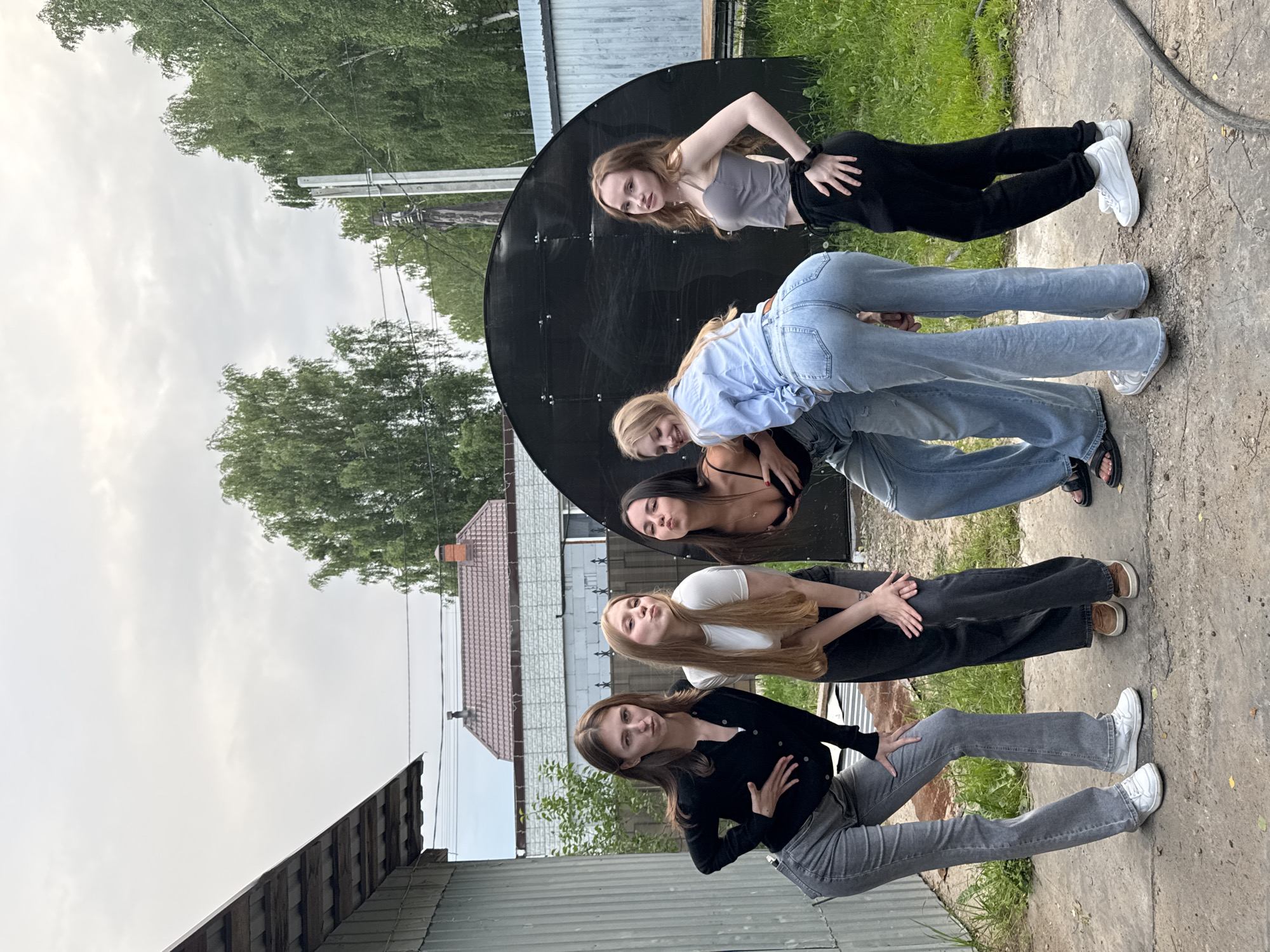 In this outdoor medium shot, six young women are posed against a large, black, circular object, possibly a satellite dish, set in a grassy, somewhat unkempt yard. The women are arranged in a staggered line, with some in the foreground and others further back. The lighting suggests it is daytime, likely late afternoon, with overcast skies and diffused natural light.
The woman at the top of the frame, on the left, has long, wavy blonde hair and is wearing a grey tank top and black pants. She is leaning back and looking towards the left. Below her, a woman with blonde hair is wearing a light blue collared shirt and jeans, and she is bending forward. Another woman with blonde hair, wearing a white t-shirt, is also bending forward, her face turned towards the viewer with puckered lips. Next to her, a woman with dark hair is wearing a black top and dark pants. Further down, a woman with dark hair is wearing a black cardigan and grey jeans, her hand on her chest. The woman at the bottom of the frame, on the right, has dark hair and is wearing a black top and grey jeans, her hand resting on her thigh.
The setting appears to be a rural or suburban area, with a building visible in the background to the left, and a wooden fence or structure to the far left. The ground is a mix of concrete and grass. The women's poses are varied, with some leaning and bending, suggesting a playful or choreographed activity. The overall mood seems casual and youthful. There is no visible text in the image. The context provided places the location as Perm, Russia.