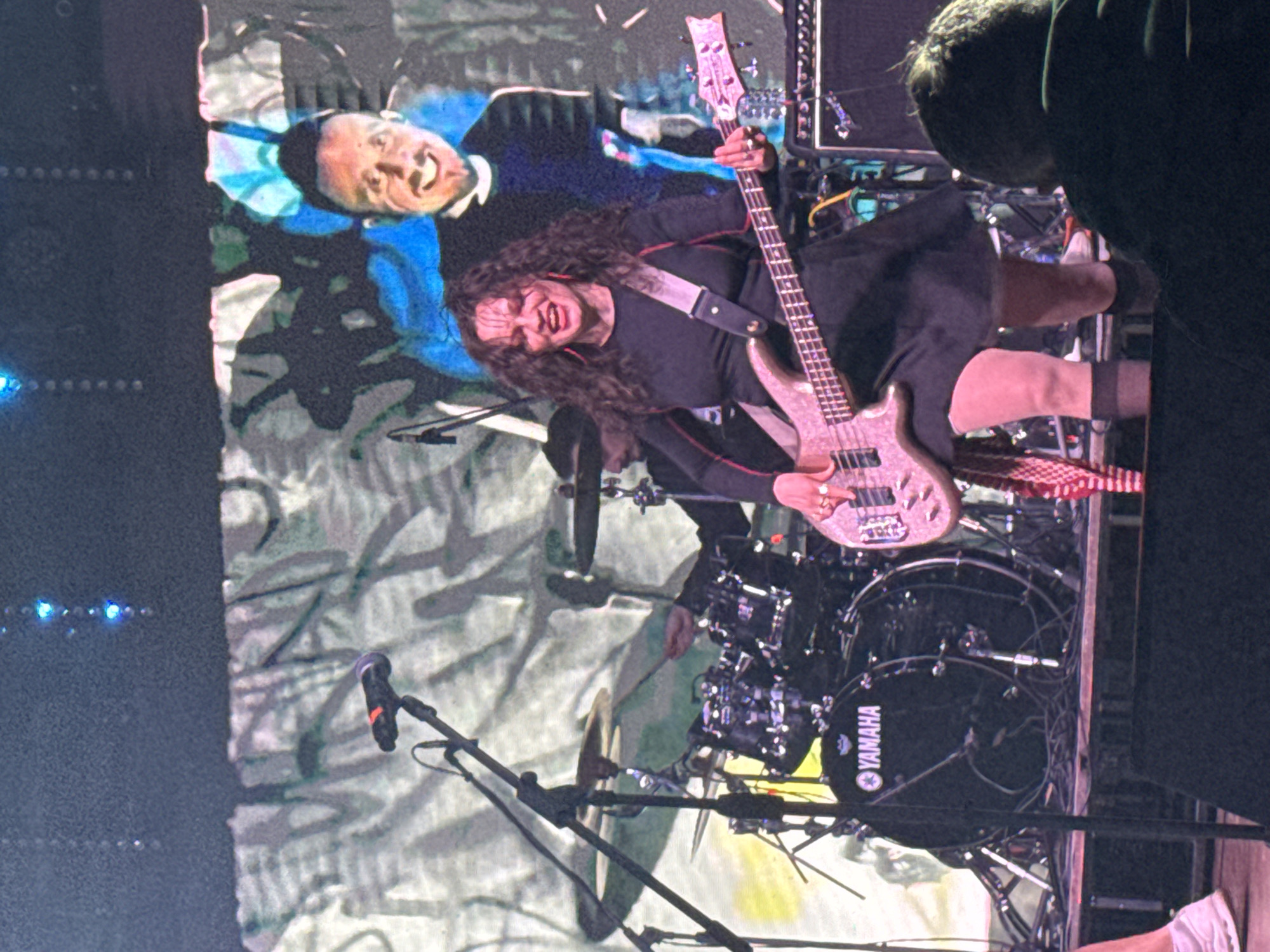 The image captures a live musical performance on a stage in Большая Кивара, Russia. A female musician with curly dark hair, dressed in a black long-sleeved dress, black sheer stockings, and light-colored shoes, stands centrally, actively playing a light-colored bass guitar. Her expression indicates intense focus or singing, with her mouth open and red earrings visible. A light-colored strap secures the bass. To the left, a male drummer, wearing a dark cap, is partially visible behind a black Yamaha drum kit, engaged in playing with drumsticks. The stage floor is dark, containing various cables and audio equipment, including a partially visible black amplifier to the right of the bassist. The backdrop is a large screen displaying abstract patterns in tones of white, grey, green, and blue, overlaid with a projected image of a smiling man with short dark hair and a beard. Overhead, blue stage lights are visible. In the bottom right foreground, the back of an audience member's head, wearing a dark garment, is visible.