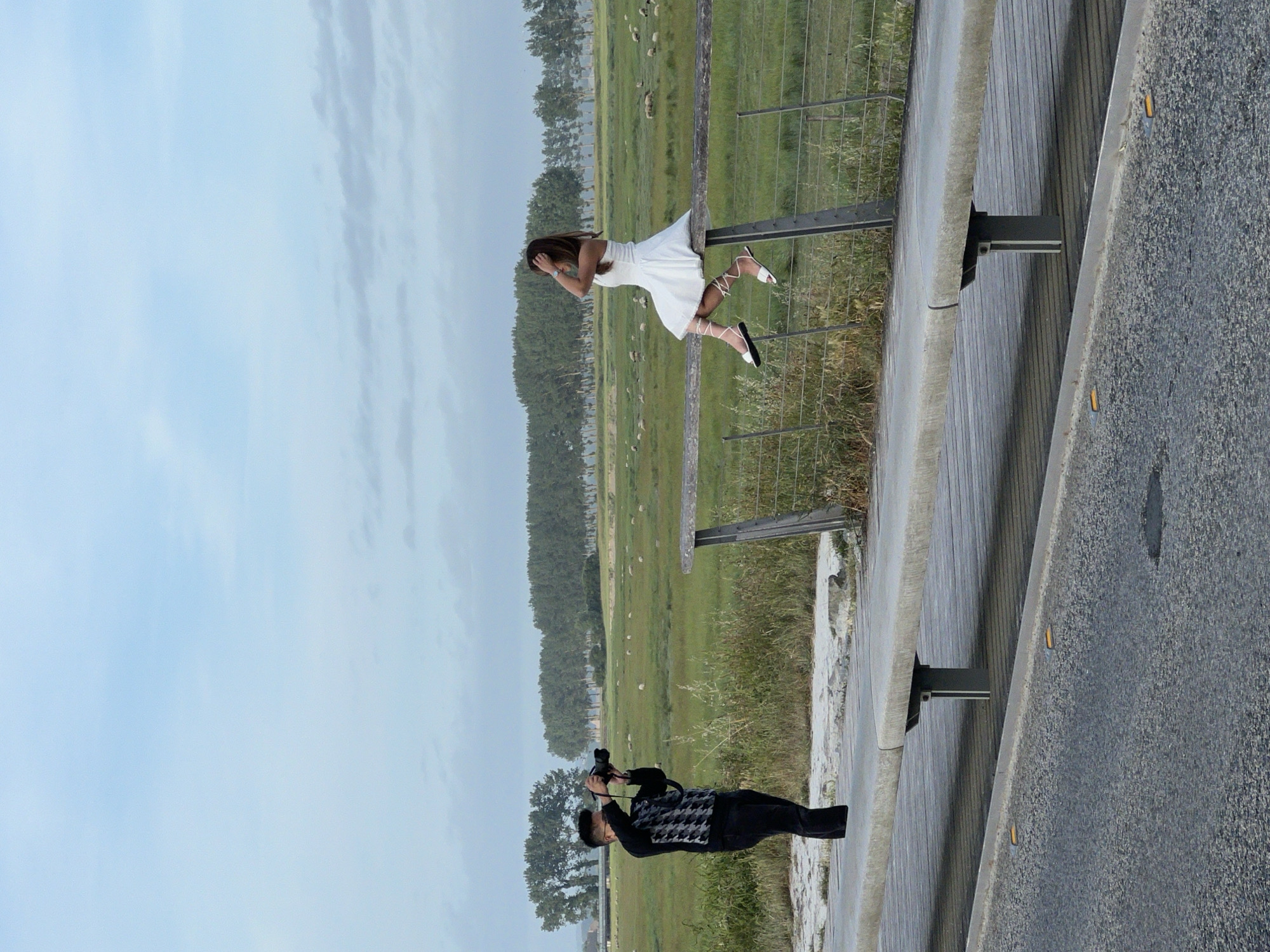 The image displays an outdoor photoshoot occurring near Le Mont-Saint-Michel, France. On the left, a man with dark hair and glasses, wearing a black and white geometric patterned sweater and dark trousers, stands on the concrete edge of an elevated walkway. He holds a black DSLR camera with a lens, pointed towards the right. On the right, a woman with long brown hair, dressed in a white sleeveless dress, sits sideways on a rustic wooden fence rail supported by grey metal posts. Her right hand is raised towards her head, touching her hair. Her legs are crossed at the ankles, and she wears white lace-up sandals with black soles. Behind the fence, a vast green field stretches to the horizon, dotted with numerous white sheep. A prominent line of tall, slender green trees forms a distant backdrop. In the lower foreground, an asphalt road features two embedded yellow reflective markers and a small dark mark. The sky is light blue with scattered diffuse grey clouds.