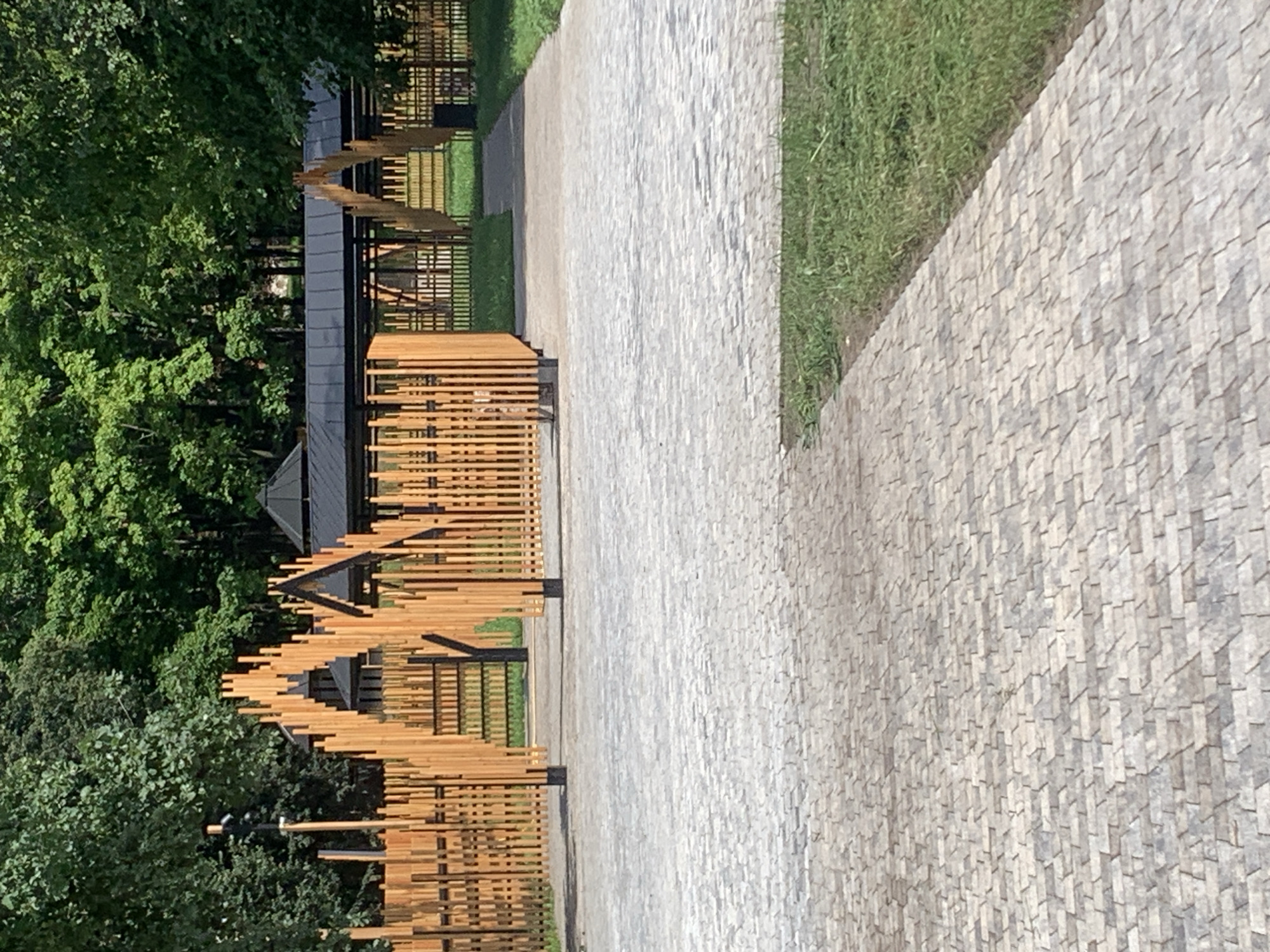 The image depicts an outdoor park area with a paved pathway in the foreground and a wooden fence structure with a covered pavilion in the background. The pathway is made of small, grey interlocking stones. Beyond the pathway, there is a green lawn area. The fence is constructed from vertical wooden planks arranged in a decorative, arched pattern. Behind the fence, a dark-colored pavilion with a sloped roof is visible. The environment is lush with mature green trees surrounding the area. There are no visible people or animals in the image. The scene appears to be in a well-maintained public space, possibly a park or recreational area.
