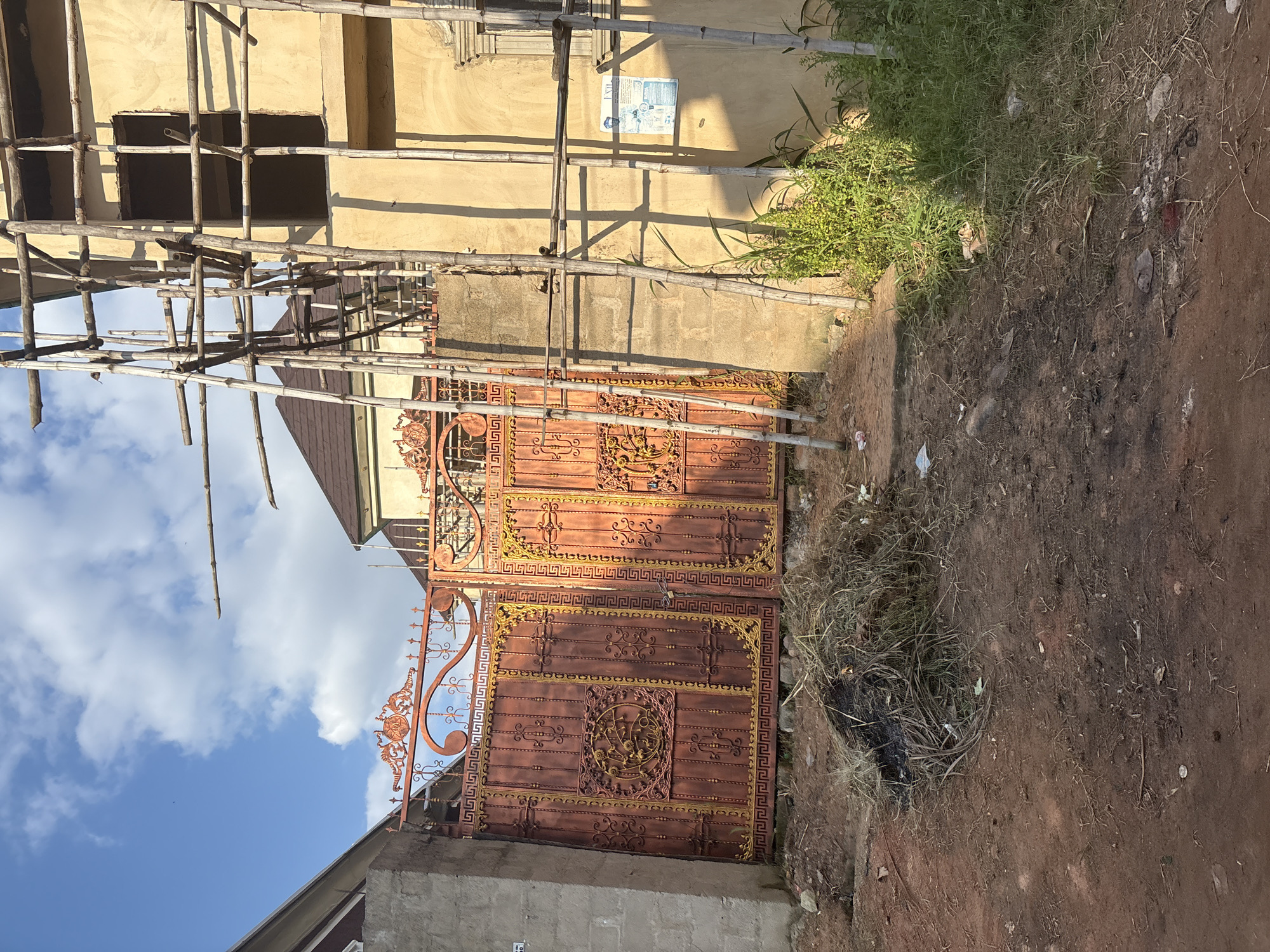 A low-angle medium shot captures a construction site with a large, ornate, copper-colored gate in the foreground, partially obscuring a building under scaffolding. The gate is adorned with intricate yellow metallic scrollwork and geometric patterns. To the right of the gate, a light yellow building is visible, covered in bamboo scaffolding that extends upwards. The building has a dark brown roof and an unfinished window opening. A poster is affixed to the building wall. A pile of dry grass and debris is located on the red soil in front of the gate. In the background, another building with a similar dark brown roof can be seen. The sky above is blue with scattered white clouds, and sunlight casts shadows across the scene. The location is identified as Obosi, Nigeria.