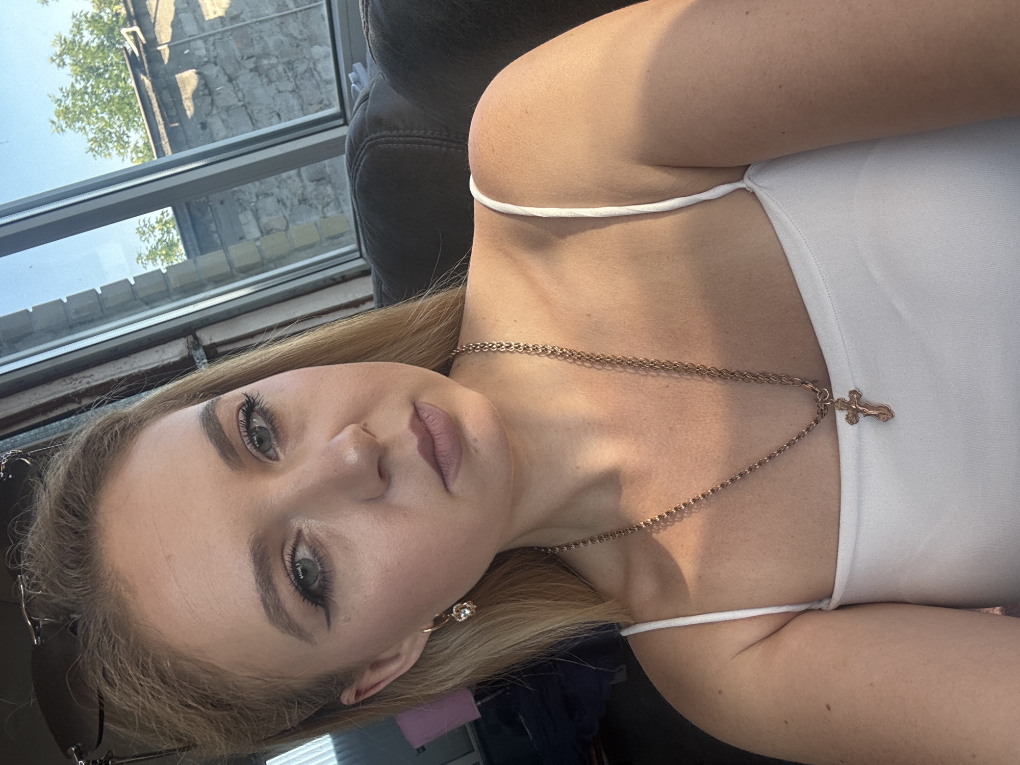 A young Caucasian woman with blonde hair, fair skin, and blue eyes is shown in a close-up, upper-body selfie taken indoors. She is wearing a white tank top and a gold chain necklace with a cross pendant. Her makeup includes winged eyeliner and neutral-toned lipstick. She is also wearing a dangle earring with a clear gem. Her hair is partially pulled back, and sunglasses are resting on top of her head. The background consists of a dark-colored couch and a window with a view of brick buildings and trees. The lighting suggests it is daytime.