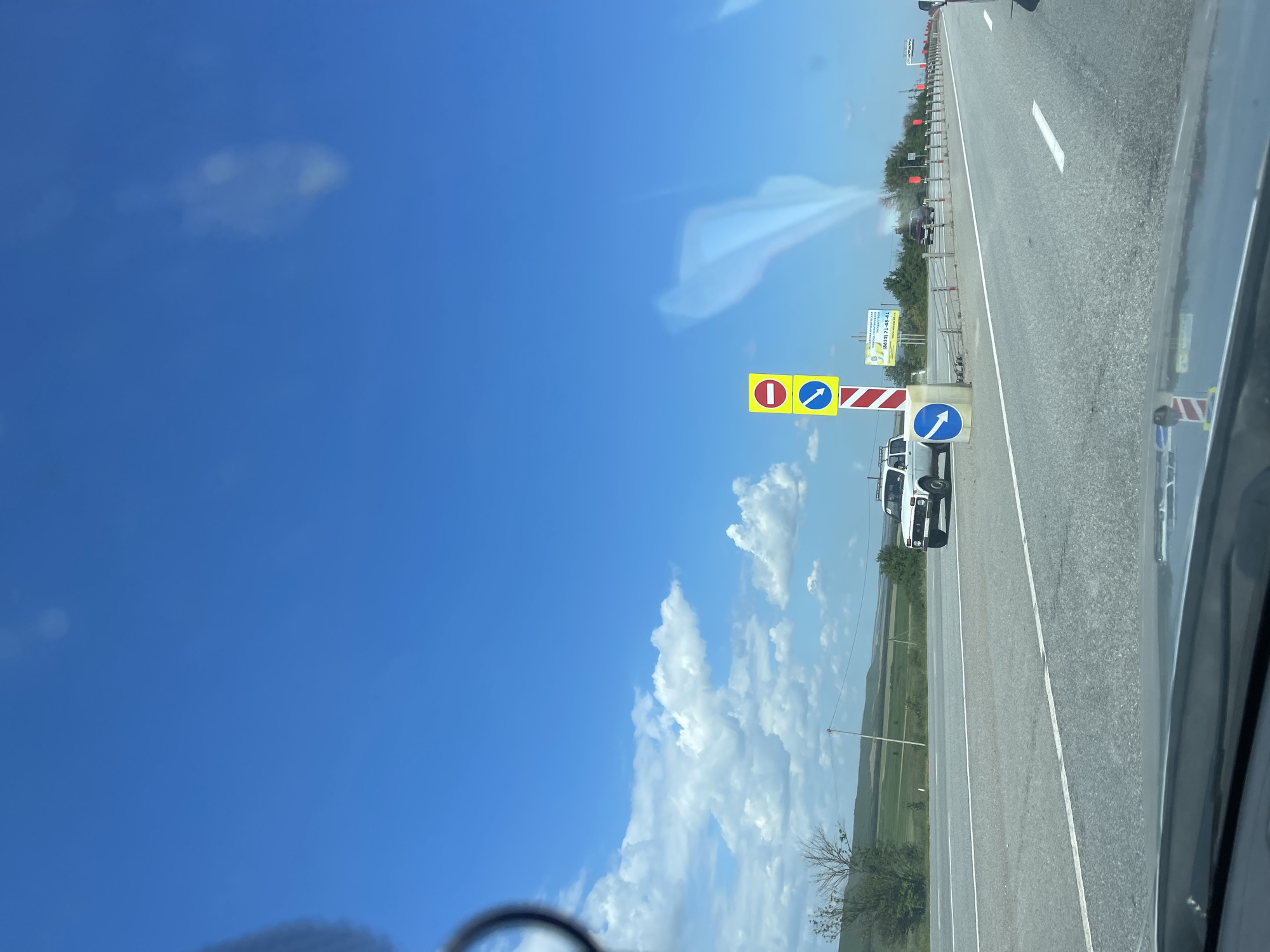 This image depicts a scene on a highway in the vicinity of Загорский, Russia. The perspective is from inside a vehicle, looking forward. The sky is predominantly blue with scattered white clouds. On the right side of the road, there are traffic signs. One sign features a red circle with a horizontal bar (prohibitory sign) and a blue square with a white arrow pointing diagonally down and to the left. Adjacent to this is a striped pole. Further down the road, a white SUV is visible in the left lane. Beyond the SUV, the landscape consists of green fields and rolling hills. The highway itself has multiple lanes separated by white lines. Other traffic signs and barriers are present along the side of the road, and a billboard with text and a phone number is visible in the distance. The overall environment suggests a daytime scene on a paved road in a rural or semi-rural area.