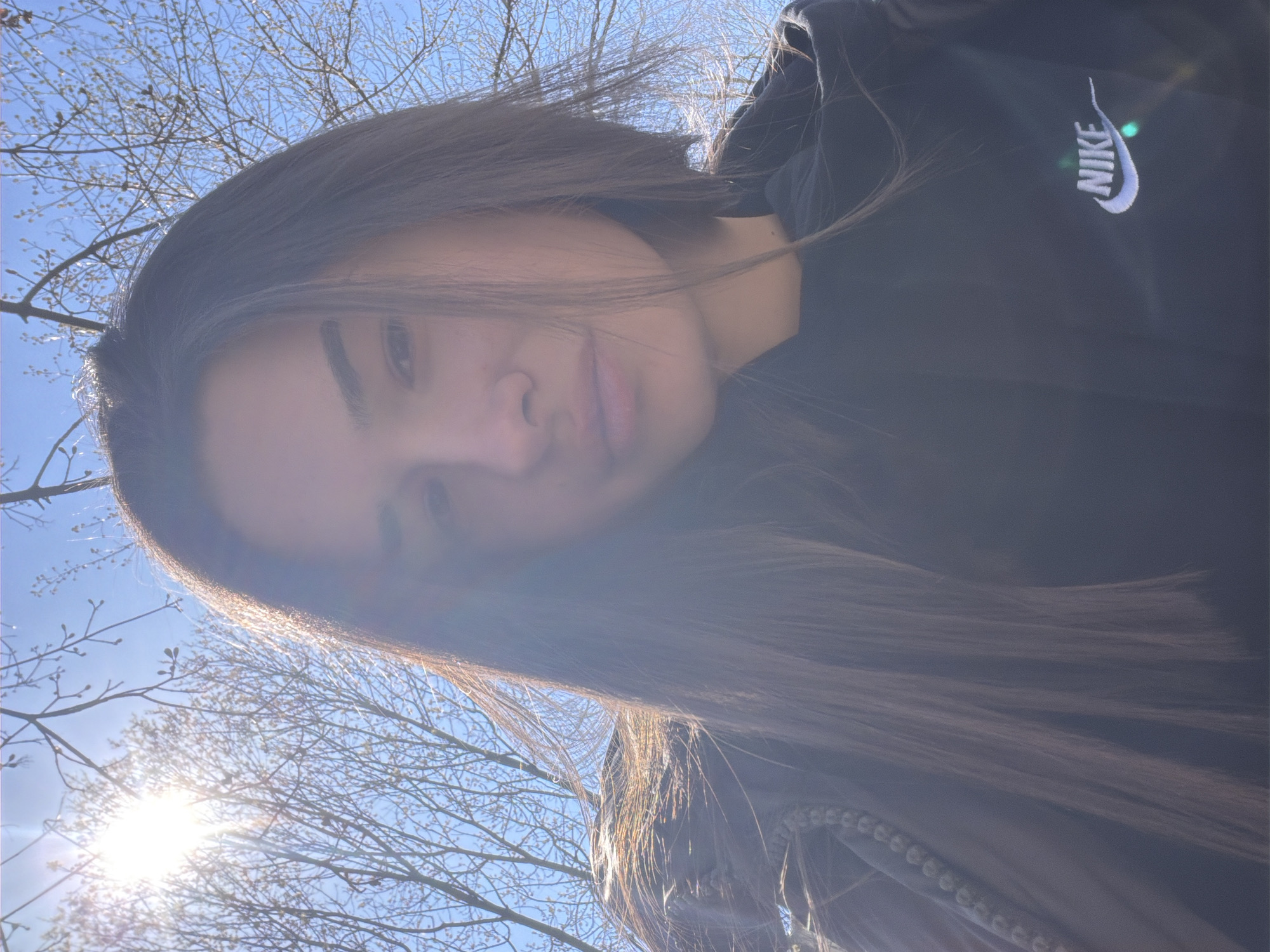 A woman with long brown hair is looking into the camera. She is wearing a black Nike hoodie. The sky is blue with bare tree branches visible in the background. The sun is shining brightly. Without more information or geographic clues within the image, I cannot provide a specific location in Sofia, Bulgaria.