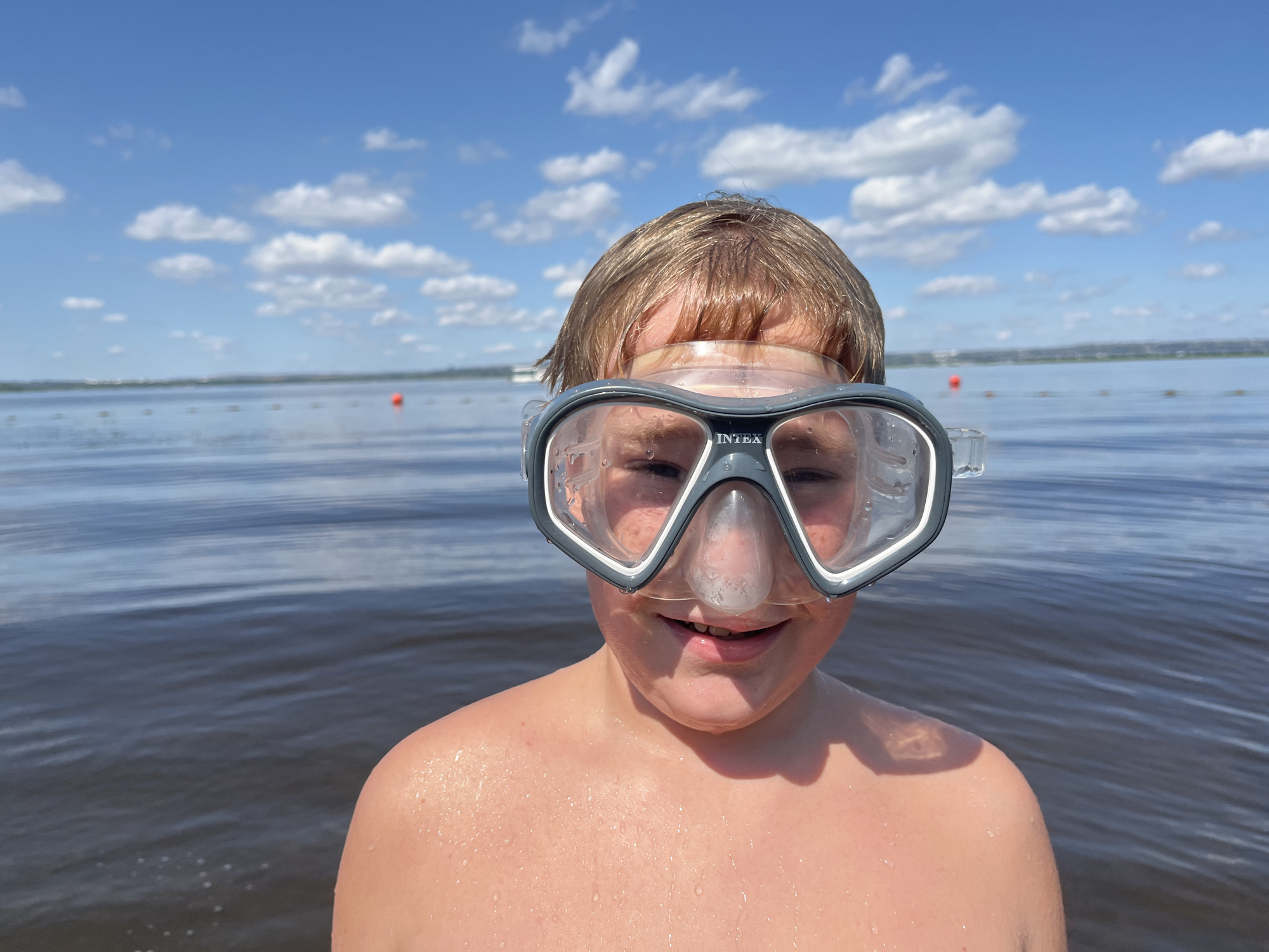 A young boy with blonde hair and a snorkel mask over his eyes smiles at the camera. He is in the water, with a body of water and a blue sky with white clouds in the background. There are orange buoys visible in the water. The scene appears to be a recreational area.