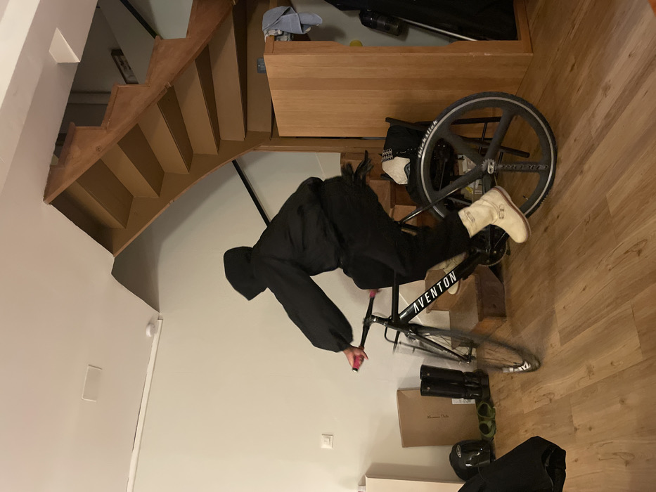 A person in a black hoodie and light-colored boots is riding a black Aventon bicycle. The bike is on a hardwood floor near the bottom of a wooden staircase. Other objects on the floor include a box, cycling shoes, and a helmet. A cabinet sits at the bottom of the staircase. The room has white walls and is brightly lit.