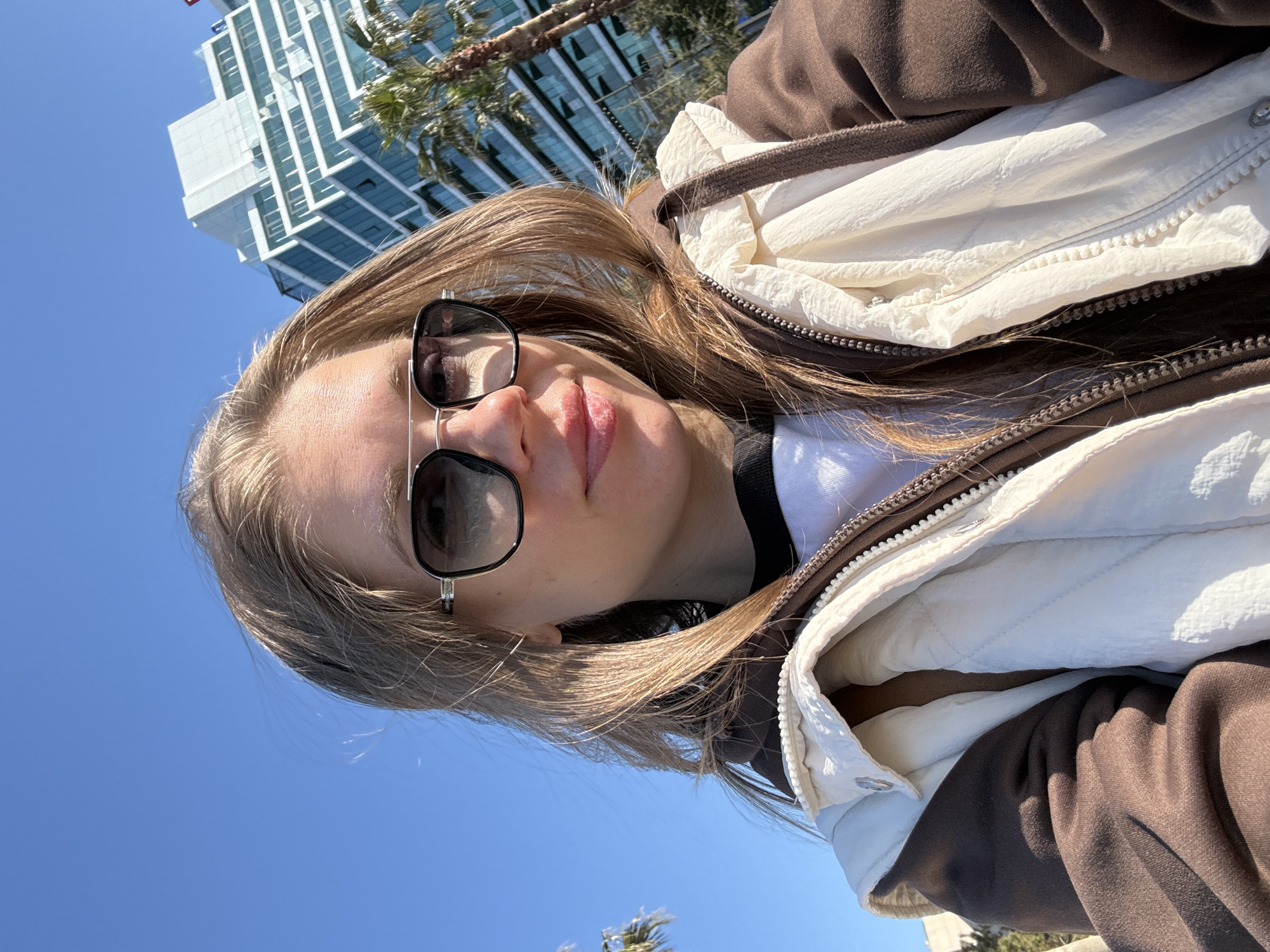 This bright, sunlit selfie is taken outdoors, featuring a young person, likely a woman, with light brown hair. She is wearing dark sunglasses that reflect the bright blue sky. Her attire consists of a white t-shirt, a brown hooded jacket, and a cream-colored quilted vest or jacket layered over the brown one. Her lips are slightly parted, with a subtle pink hue.
The background shows a clear, vibrant blue sky dominating the upper portion of the frame. Modern, glass-fronted high-rise buildings are visible in the distance, angled due to the camera's orientation. Several palm trees are scattered throughout the background, indicating a warm, possibly coastal, urban environment. The bright lighting suggests it is daytime with clear, sunny weather. The overall mood is casual and relaxed. There is no visible text in the image. This scene is consistent with the provided location context of Batumi, Georgia, a city known for its modern architecture and coastal setting.