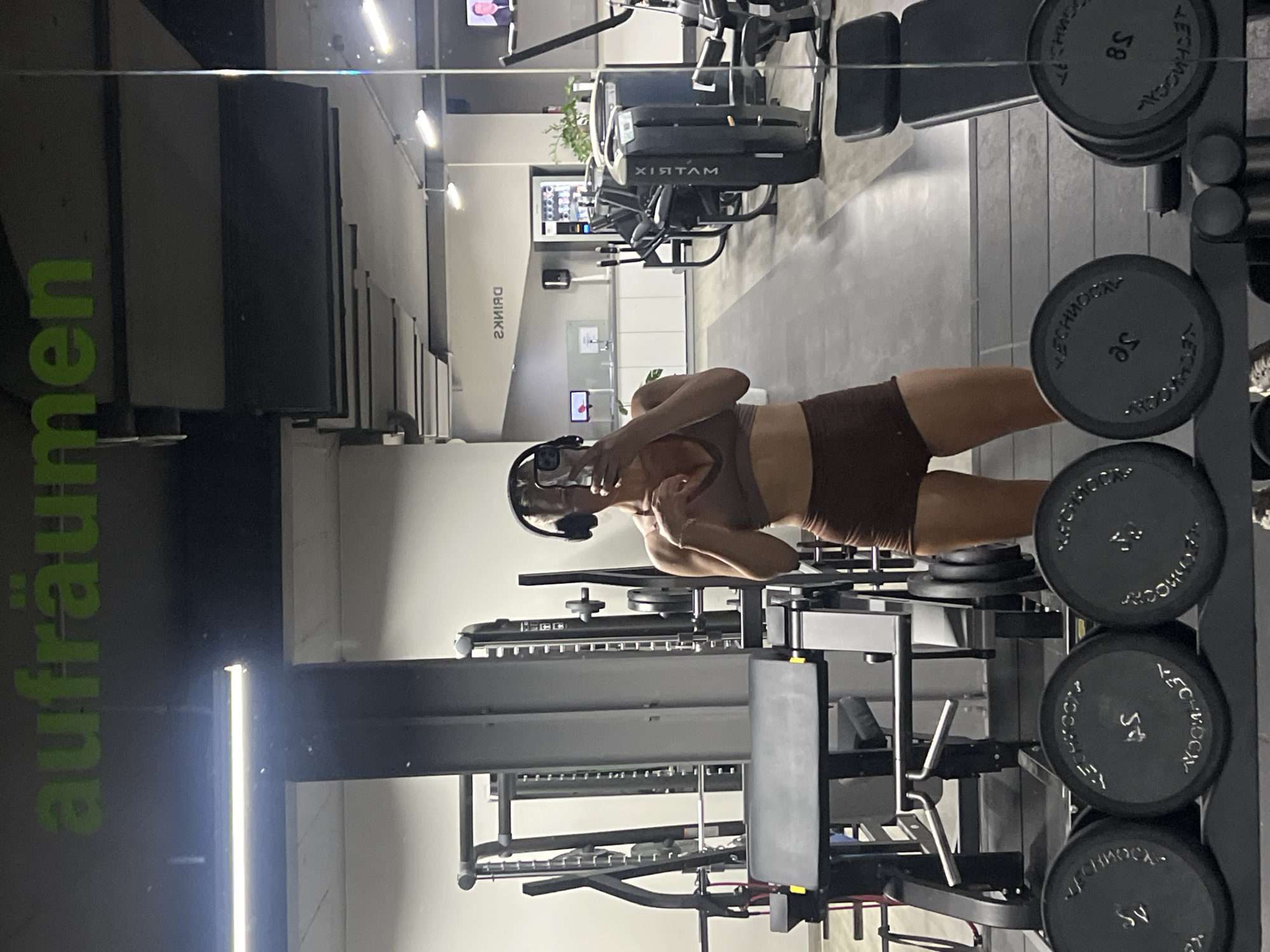 A full-length mirror selfie shows a woman in a gym in Altötting, Germany. The woman, wearing a brown sports bra and brown shorts, has headphones on and is holding her phone to take the picture. In the background, various gym equipment is visible, including weight racks with plates, a leg press machine, and other exercise machines. The gym has a modern design with metal accents and overhead lighting. A sign on the wall reads "AUFRÄUMEN," which translates to "tidying up" or "cleaning up" in German, and another sign in the reflection indicates "DRINKS." The overall atmosphere suggests a well-equipped fitness facility.
