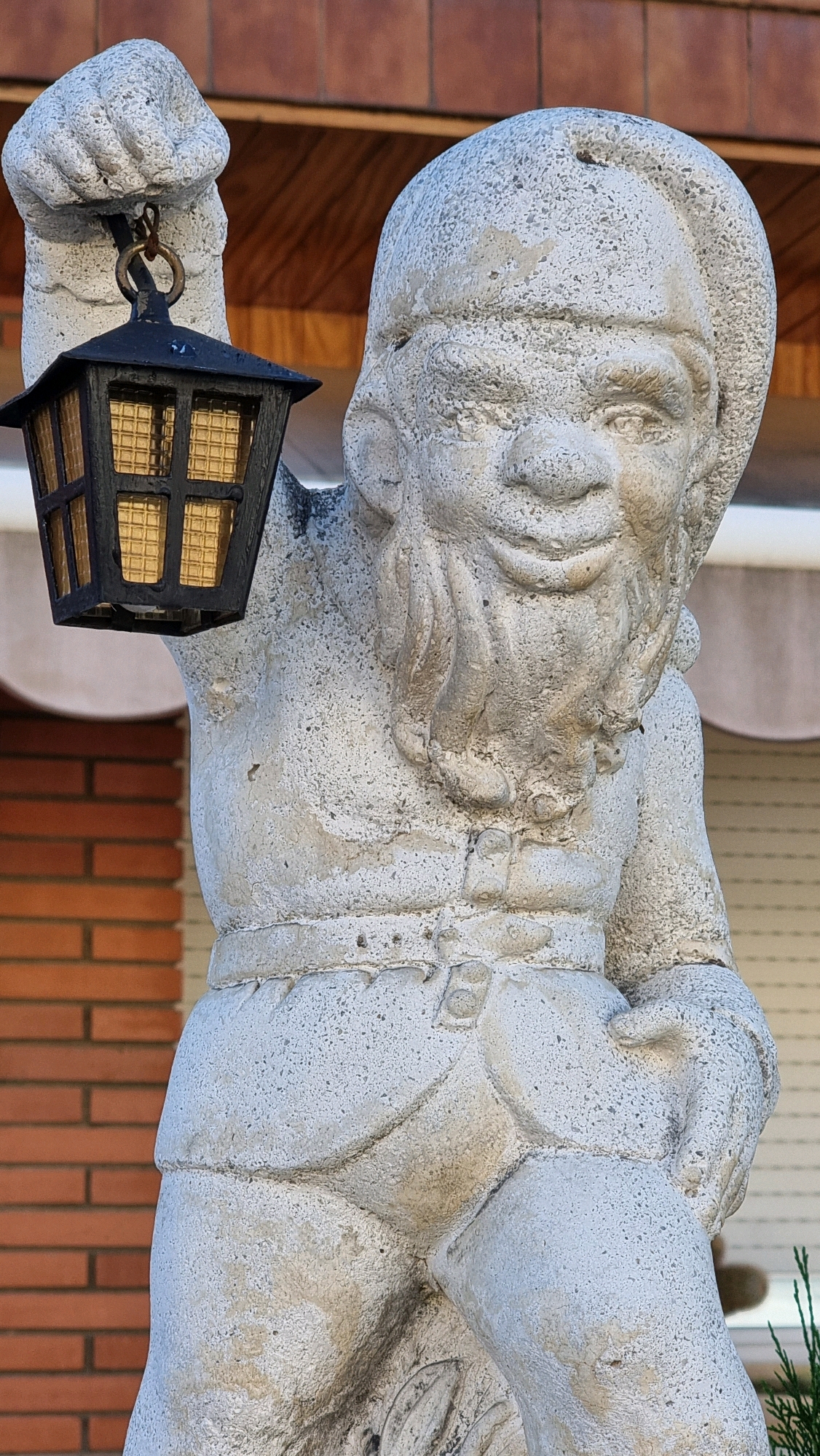 This is a close-up, eye-level, outdoor photograph of a weathered, white stone gnome statue holding a black lantern with amber glass panes in its raised right hand. The statue is detailed, with a hat, a beard, and clothing depicted. The background shows a section of red brick wall on the left and a beige awning or shutter on the right. The statue appears to be located in the town of Sant Joan Les Fonts, Spain, likely in a garden or on a property.