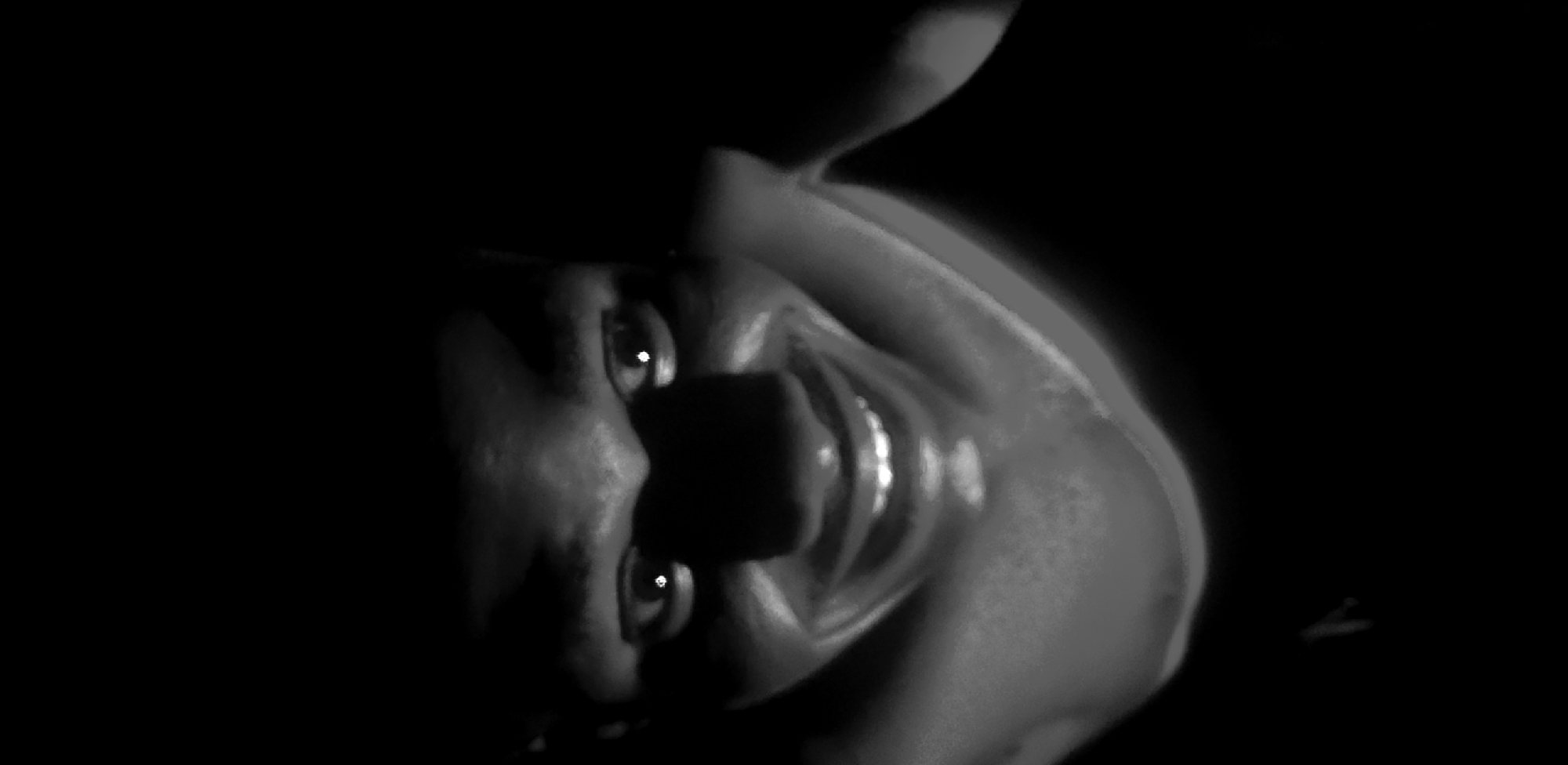 The image shows a close-up, black and white shot of a person's face, lit from below. The lighting creates harsh shadows, emphasizing the contours of the face and giving it a sinister appearance. The person has wide eyes with bright reflections, a prominent nose, and a wide, toothy smile. The background is entirely black, suggesting a dark environment with no discernible features. There is no information in the image to indicate a location in Narayi, Nigeria, or any urban setting.