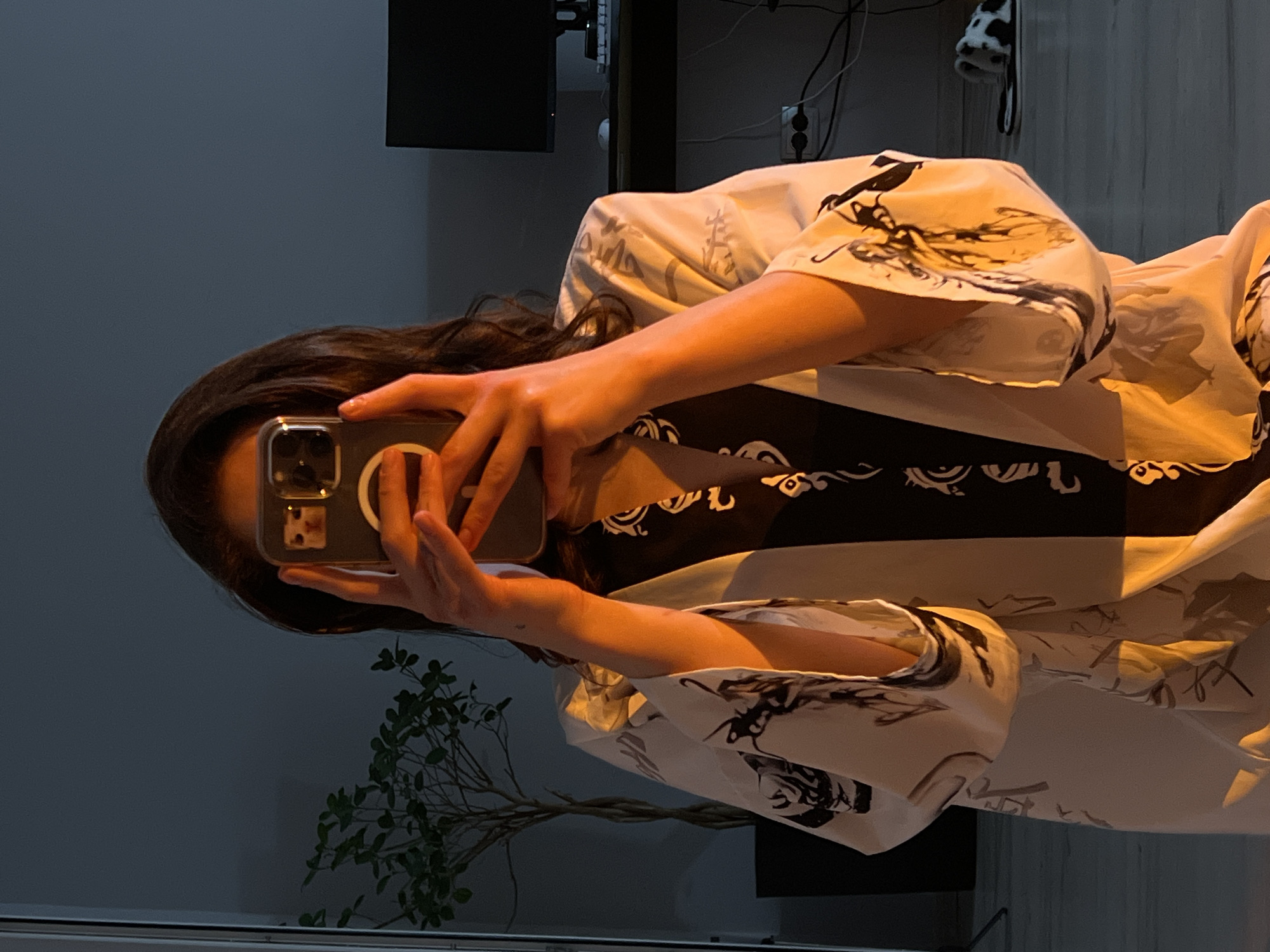 A person is taking a mirror selfie in a dimly lit room, likely an apartment. The person is holding a smartphone with a case featuring a cat sticker, obscuring their face. They are wearing a dark patterned top layered under a white robe with black ink-like designs. The room appears to be a living space or bedroom, with a potted plant visible to the left and a desk with a computer monitor and peripherals in the background to the right. A pair of fuzzy slippers with a cow-like pattern are on the floor to the far right. The lighting is warm and low, casting dramatic shadows. There is no specific indication of the location being in Ternopil, Ukraine.
