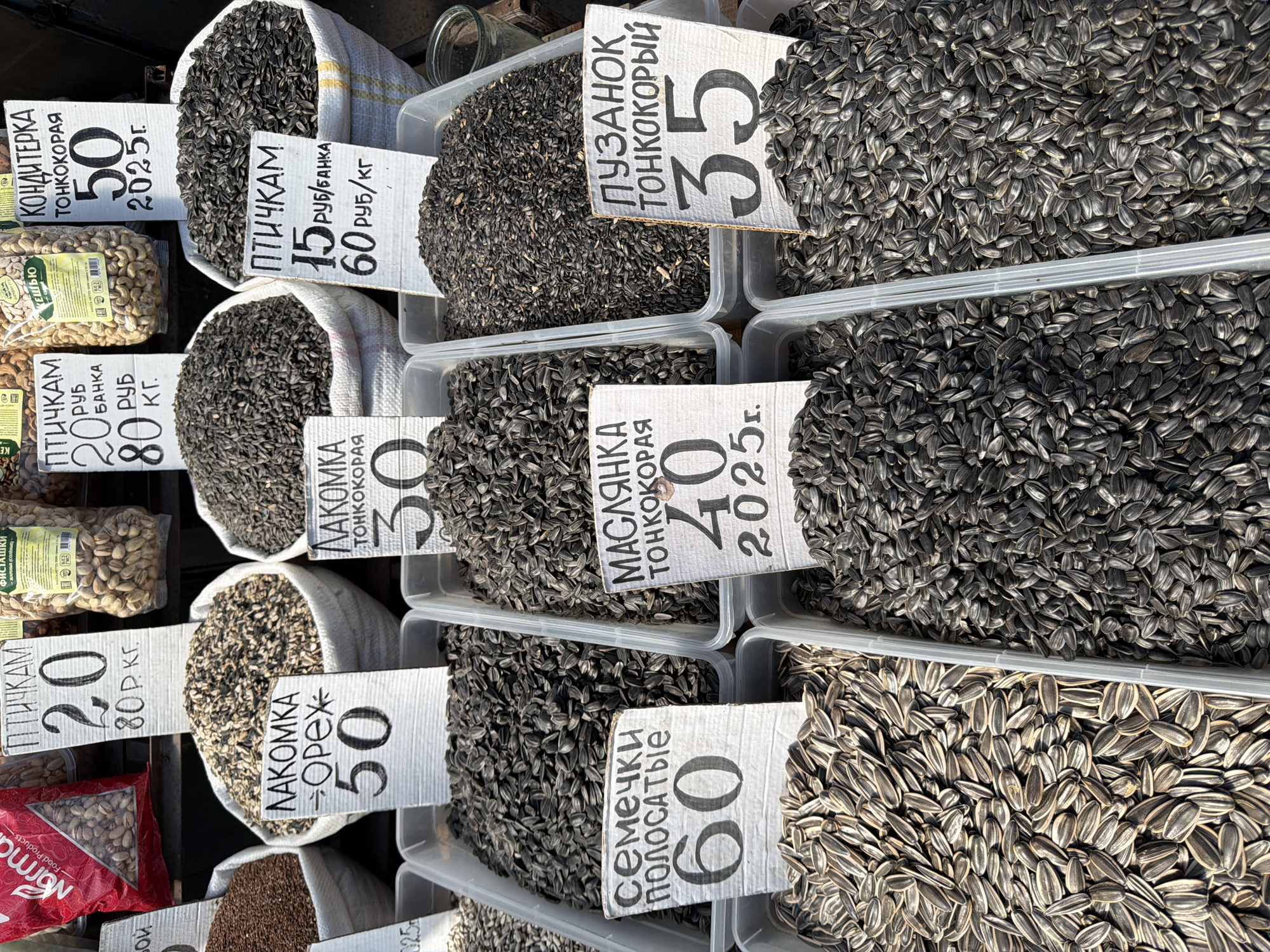 The image displays a street market stall in Taganrog, Russia, showcasing a variety of bulk edible seeds. The stall features several large white sacks and plastic containers filled with different types of seeds, including sunflower seeds in shells and shelled varieties. Each container or sack has a handwritten price tag displayed prominently.
The price tags include product names and prices in Russian rubles. For example, one tag reads "ЛАКОМКА ОРЕХ 50" (Delicious Nut 50), another "СЕМЕЧКИ ПОЛОСАТЫЕ 60" (Striped Seeds 60), and another "МАСЛЯНКА ТОНКОКОРАЯ 40" (Oily Thin-shelled 40). There are also tags indicating prices for bird feed, such as "ПТИЧКАМ 20 руб. банка 80 руб. кг." (For Birds 20 rubles per jar, 80 rubles per kg). Another tag reads "ПУЗАНЮК ТОНКОКОРЫЙ 35" (Fatty Thin-shelled 35). The prices are denominated in Russian rubles, with some indicated as per kilogram (кг) or per jar (банка). Some tags also display a year, such as "2025 г." (2025).
The setting appears to be outdoors, likely a market, with the natural light illuminating the scene. The background is somewhat blurred, suggesting a shallow depth of field, focusing attention on the products for sale. There are no people visible in the immediate foreground or interacting with the products. The overall impression is one of a typical market scene, focused on the sale of food items.