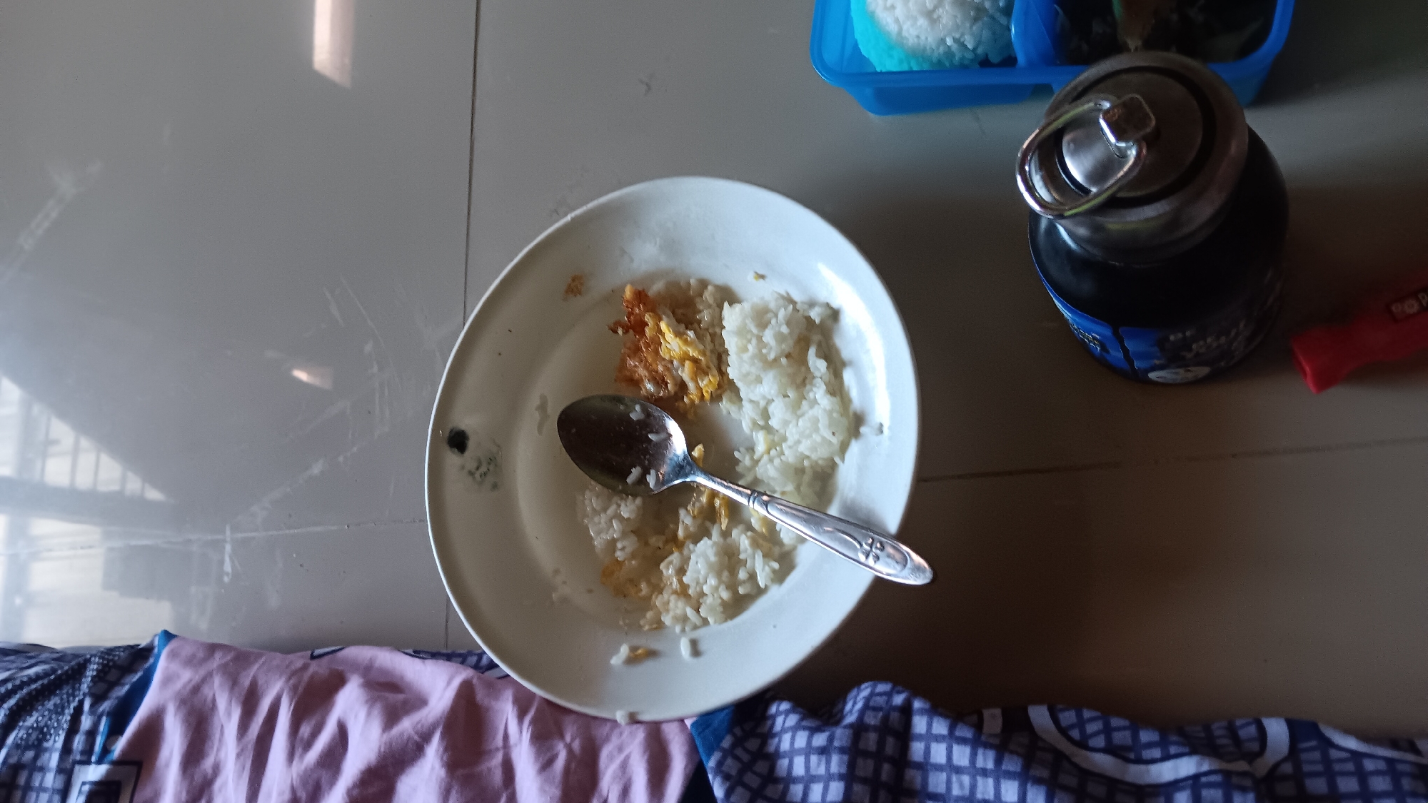 In the city of Sonobekel, Indonesia, a close-up, overhead shot captures a partially eaten meal of rice and scrambled eggs on a white plate, with a silver spoon resting in the food. The plate is situated on a tiled floor, adjacent to a folded pink and blue patterned fabric. To the right of the plate, a blue plastic container holds a portion of white rice. Further to the right, a dark water bottle with a silver ring on its cap is visible, and a red object, possibly a marker or tool, lies nearby. The environment appears to be indoors, with overhead lighting reflecting off the tiled floor.