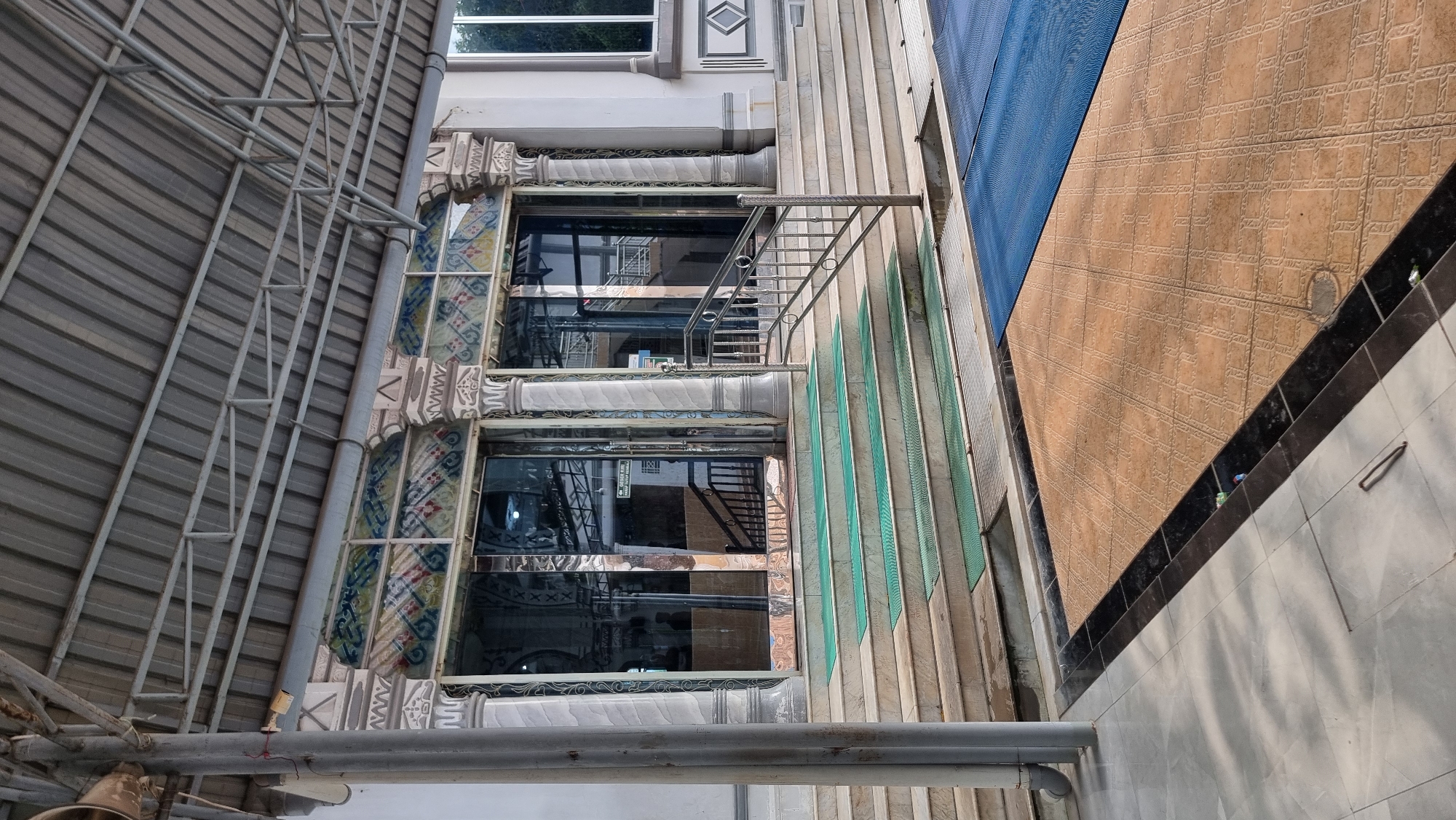 The image depicts the entrance of a mosque in Keudekruenggeukueh, Indonesia. A metal roof supported by metal beams extends outwards, providing shade over the entrance area. The entrance itself features a set of wide marble stairs leading up to glass doors. Green mats are placed on some of the steps. Decorative columns with intricate carvings flank the entrance, and a patterned frieze runs above the doors. To the right of the stairs, a silver metal railing guides visitors. The foreground shows tiled flooring, with a blue mat and a tan tiled area visible. The overall scene is an exterior view of a religious building, suggesting a place of worship accessible to the public.