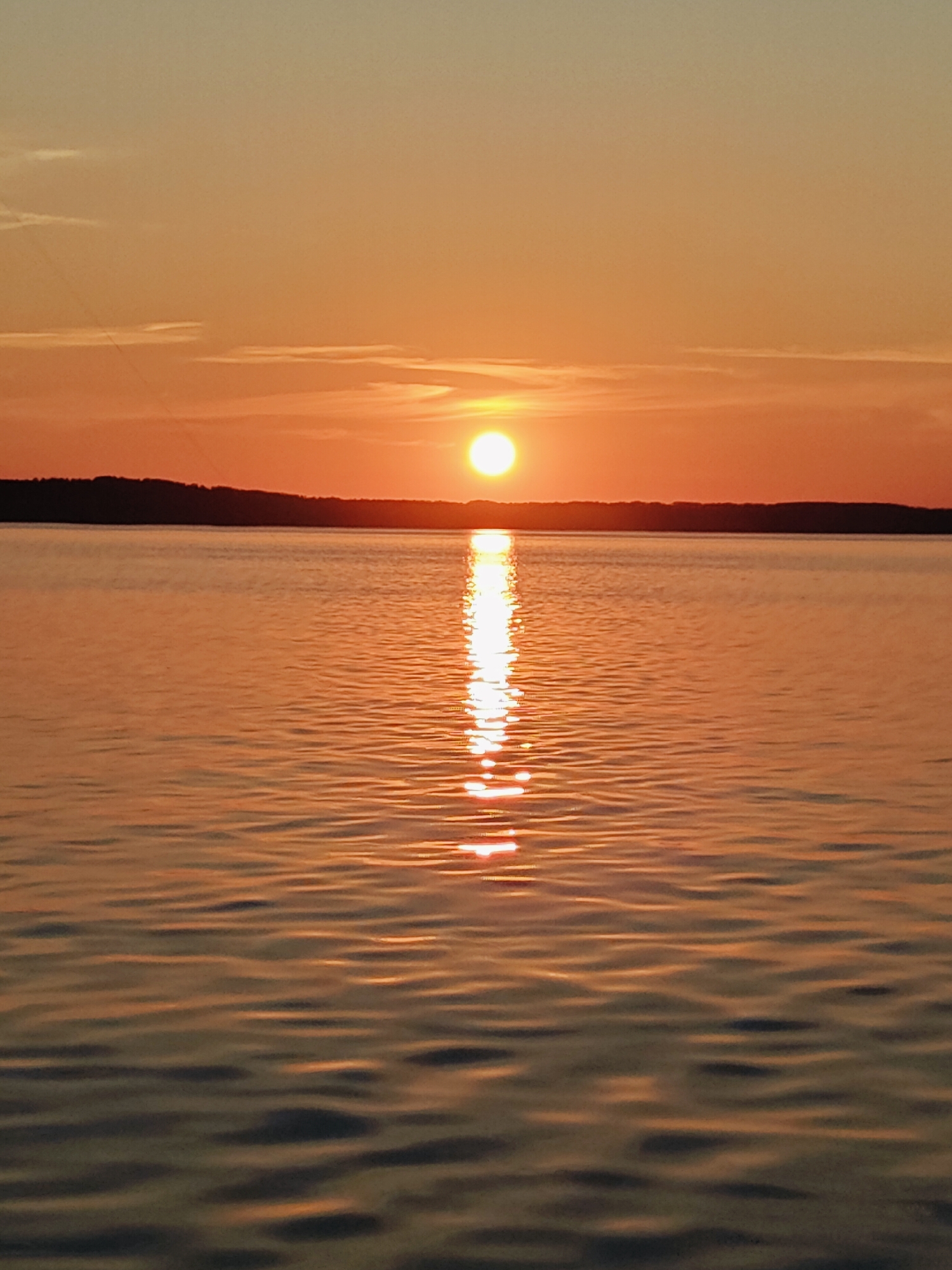 The image shows a tranquil sunset over a large body of water, likely the Kama River in Chaykovsky, Russia. The sun, a bright, glowing orb, is positioned just above the horizon, casting a warm, orange hue across the sky and reflecting a shimmering path on the water's surface. Gentle ripples disturb the otherwise calm water. In the distance, a dark, tree-lined shore forms a silhouette against the vibrant sky. The overall atmosphere is peaceful and serene. No people or specific objects are clearly visible beyond the natural elements of the scene.