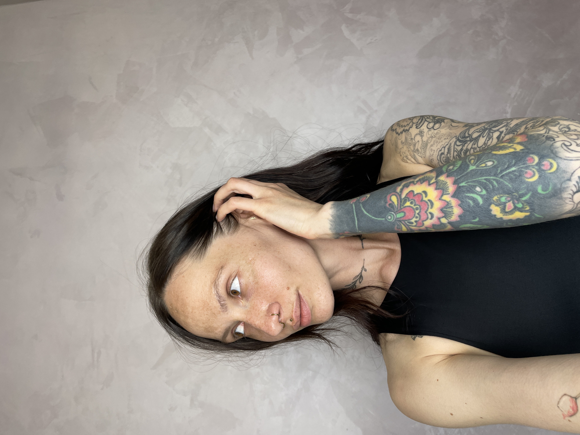 A woman with dark hair and tattoos is posing in front of a gray wall. She is wearing a black tank top. Her left arm is raised, with her hand touching her hair. She has a nose ring. There is no indication of the location of Izhevsk, Russia.