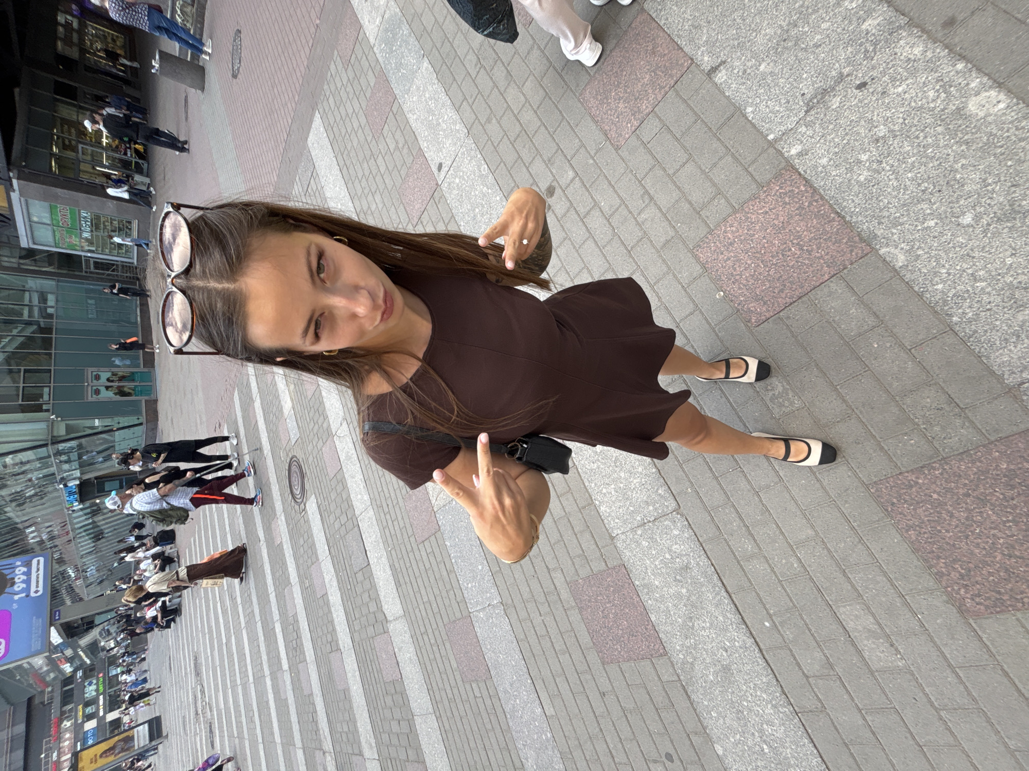 The image depicts a young adult female posing for a photograph on a paved pedestrian area in Saint Petersburg, Russia, with a busy urban background.
**Location:** The scene is identifiable as an area in Saint Petersburg, Russia, specifically around Nevsky Prospekt and Ploshchad Vosstaniya, suggested by the prominent blue billboard on a building displaying "1999" and the Russian text "УЛЫБАЕМСЯ ЧАЩЕ" (Smile more often). The modern glass architecture and the style of the pedestrian crossing are consistent with this commercial district.
**Main Subject:** In the immediate foreground and center, a light-skinned female with long brown hair, parted in the middle, is looking directly at the camera. She has a slightly puckered lip expression. She is wearing a dark brown, short-sleeved mini-dress and black and beige Mary Jane style flat shoes. Sunglasses with brown frames and reflective lenses are pushed up onto her head, reflecting clouds. A small black shoulder bag hangs on her left side. She is performing a peace sign gesture with both hands; her left hand, wearing a ring on the ring finger and light pink nail polish, is raised with the index and middle fingers extended in a V-shape. Her right hand is similarly gesturing.
**Background:** The background features a multitude of individuals walking and standing. A person in a long brown skirt and light top walks from right to left, carrying a bag. To the right, a male figure wearing a white baseball cap, a grey backpack, and dark pants with red stripes walks left to right, using a walking stick. He is next to a woman dressed in black. Further back, numerous people are visible, some sitting on benches along the street. The ground consists of grey rectangular pavers interspersed with larger reddish-brown granite sections. A light and dark grey striped pedestrian crossing (zebra crossing) is visible in the mid-ground. Buildings with large, reflective glass facades dominate the background, displaying various signs, including "ОТО," "ЦВЕТЫ" (Flowers), and "КУБИКА." The scene is brightly lit by natural daylight.