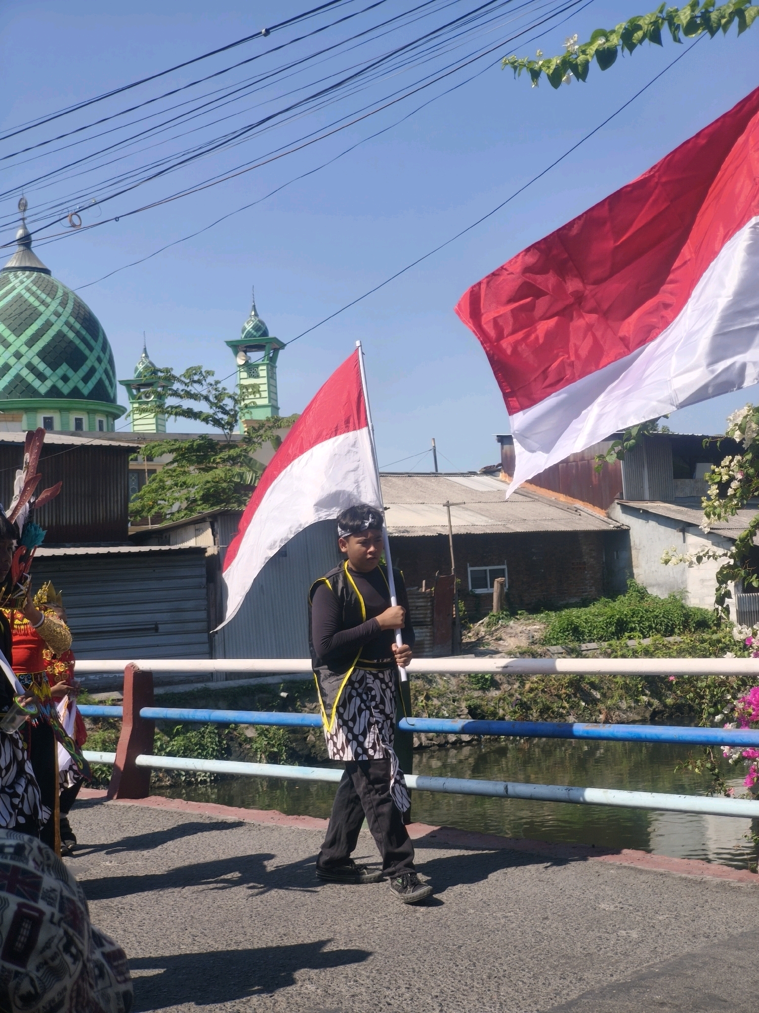 The image depicts a sunny outdoor scene on a bridge in Kepuh Kiriman, Indonesia. A young person, dressed in traditional attire including a black long-sleeved shirt, a patterned sarong-like garment, and a black headscarf, walks while holding the Indonesian flag. The flag is large and prominently displayed, with its red and white colors stretching across the frame. Behind the flag, two green-domed mosques with minarets are visible, indicating a religious and cultural setting. Several buildings with corrugated metal roofs are also present. A river flows beneath the bridge, bordered by lush greenery. Other individuals, also in traditional clothing, are partially visible in the foreground and to the left, suggesting a procession or cultural event is taking place. The sky is clear and blue, with overhead power lines contributing to the urban or semi-urban environment.