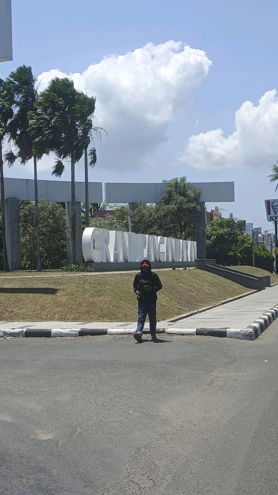 The image shows a man in a black hooded sweatshirt and blue jeans walking on a sidewalk. Behind him, large white letters spell out "GRAND PANGANDARAN," indicating the location in Pangandaran, Indonesia. Palm trees sway in the background under a partly cloudy blue sky. The man is holding a smartphone in his right hand. A KFC sign is visible on the right side of the frame. The scene appears to be a public area with paved roads and grassy slopes.