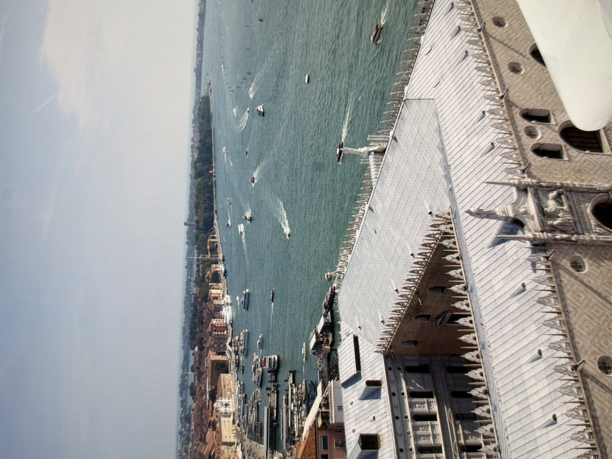 The image is an aerial view of a city with canals and numerous boats. In the foreground, there are two ornate buildings with white roofs and decorative architectural elements. The buildings have a light brown, textured facade with arched windows and intricate detailing along the roofline.
The middle ground shows a wide canal filled with various types of boats, including speedboats leaving wakes and larger passenger vessels. The water is a deep teal color. Along the edges of the canal are more buildings, some appearing older with terracotta roofs, and others more modern. There are also docks and piers with more boats docked.
The background reveals a cityscape with a dense collection of buildings stretching along the coastline. Beyond the city, a vast body of water extends to the horizon under a clear, pale blue sky. The overall impression is of a historic European city with a prominent canal system.
The scene is not located in Kyiv, Ukraine. This appears to be a depiction of Venice, Italy, given the distinctive architecture, canals, and boats.