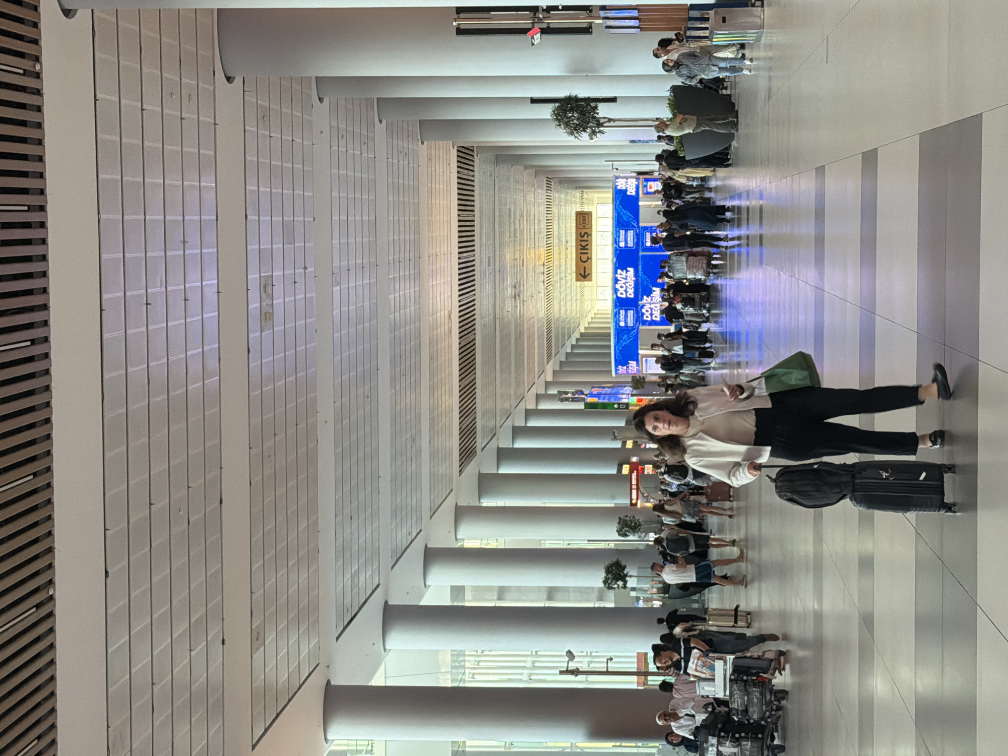 The image depicts a large, modern airport concourse in Arnavutköy, Turkey. A woman is walking towards the camera in the foreground, pulling a black suitcase and carrying a green tote bag. She is wearing a white jacket, a light-colored top, and black pants. Her right hand is holding a mobile phone. The concourse features a high ceiling with a distinctive slatted wooden upper section and a grid of illuminated panels below. Numerous large, cylindrical white pillars line the walkway, creating a sense of depth and scale. In the mid-ground, other travelers are visible, some with luggage, and a row of shops and service counters can be seen. A yellow directional sign with Turkish text "← ÇIKIŞ" (Exit) is prominently displayed. The floor is tiled with a pattern of grey and white squares. The overall atmosphere is that of a busy, functional transportation hub.