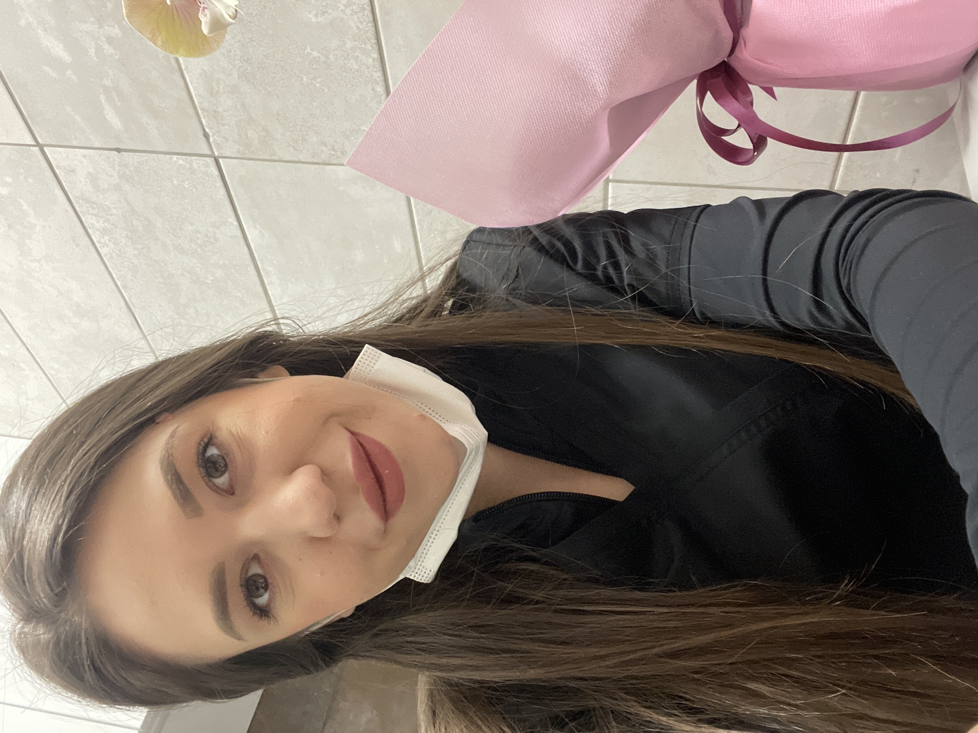 A young woman poses for a selfie indoors. She is wearing a black top and a white surgical mask pulled down below her mouth. She has long, brown hair and brown eyes. She is holding a bouquet of flowers wrapped in pink paper with a maroon ribbon. The background is a white tiled wall. The location appears to be inside a building; based on the information provided, this is in Sofia, Bulgaria.