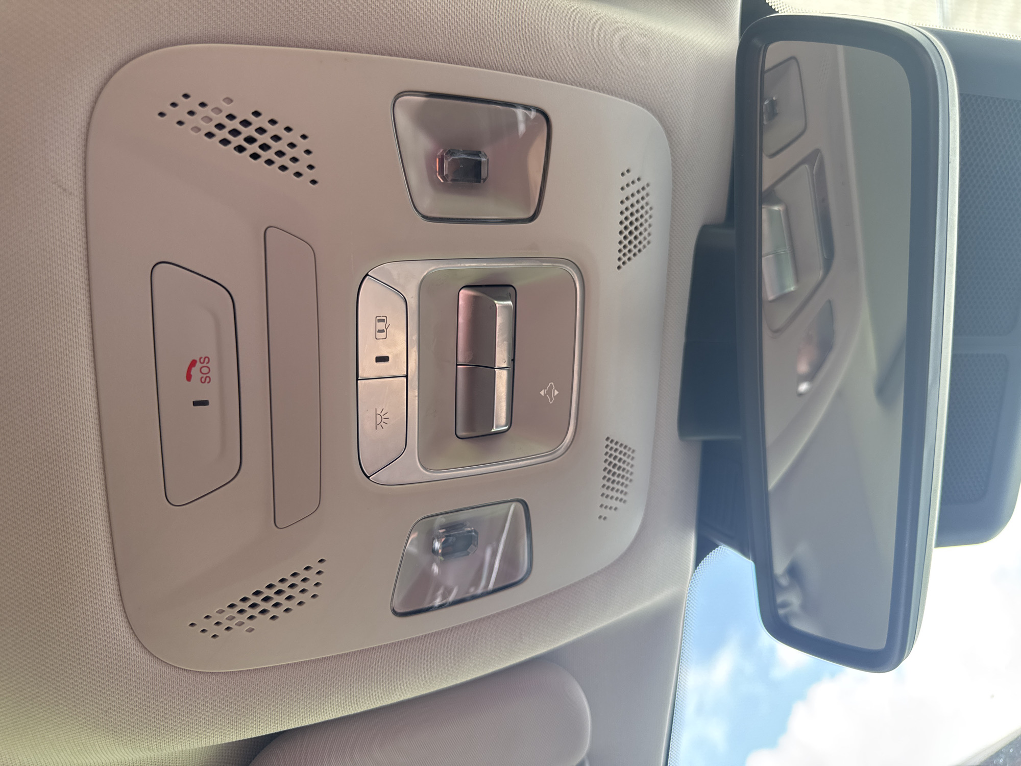 The image shows a close-up, interior view of a car's overhead console and rearview mirror, taken from the perspective of a rear passenger. The console is beige and features several buttons and lights. In the center of the console, there is a rectangular area with two prominent silver buttons, one above the other. To the left of these buttons, there is a small, rectangular light fixture with a clear cover. To the right, there is another light fixture with a clear cover, and inside, a red light is visible. Above these, there is a larger rectangular panel with the word "SOS" and a red emergency call icon. On either side of the "SOS" panel are grilles, likely for speakers or ventilation. Below the console, the car's rearview mirror is visible, reflecting the overhead console and the car's interior. The mirror itself is framed in black. Through the windshield, which is reflected in the mirror and also visible at the top of the image, a blue sky with white clouds can be seen. The overall scene suggests a modern car interior. The location of Nikolaevka, Russia, is not depicted in the image, as it only shows the interior of a vehicle.