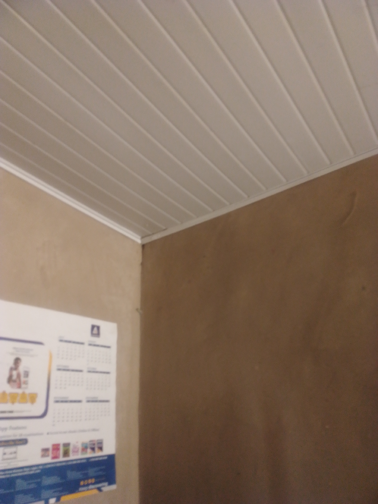 This is a medium-angle, indoor shot of a room, likely in Jauro Yinu, Nigeria. The primary focus is on a white, slatted ceiling and a brown wall. On the left side of the image, a portion of a poster or calendar is visible, mounted on the wall. The poster features a grid that appears to be a calendar with months such as "OCTOBER 2023" visible. There are also some small images of books and what looks like a person in a drawing on the poster. The wall is a neutral brown color. The ceiling is made of long, white, rectangular panels with distinct grooves between them, creating a ribbed effect. The lighting suggests it is daytime, but the interior setting limits specific cues about the time of day. There are no people or overt activities depicted, suggesting a static or unoccupied scene. The overall impression is a simple, functional interior space.