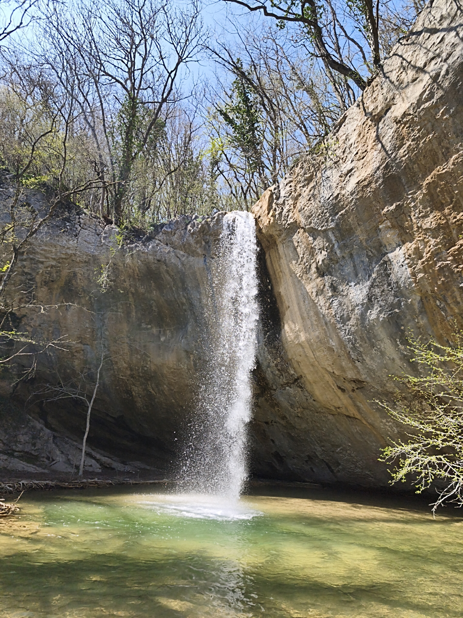 A waterfall cascades into a clear, green-tinted pool at the base of a rocky cliff, flanked by trees. The scene is located in the Орлиновский округ, Ukraine.
