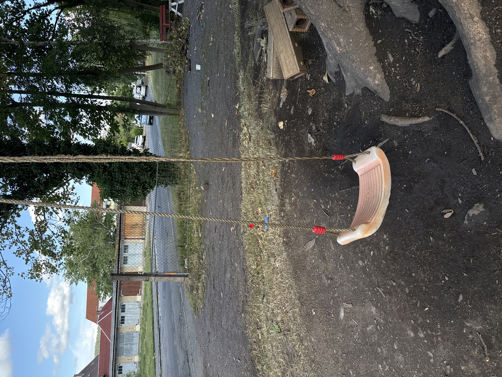 The image depicts an outdoor scene with a swing set prominently featured in the foreground. The swing has a light-colored plastic seat and thick rope ropes with red plastic attachments. The ground beneath the swing is dark, loose soil with some scattered leaves and small branches. To the right of the swing, large tree roots are visible, partially covered by the soil.
In the mid-ground, a chain-link fence runs horizontally across the image, separating the swing area from a paved road and a grassy area beyond. Several garages with various colored doors are visible along the road. Behind the garages, buildings with red roofs can be seen. Further in the background, trees with green foliage line the area, and a few cars are parked on the road. On the right side of the mid-ground, there is a wooden bench with a white table.
The sky above is blue with scattered white clouds. The overall scene suggests a somewhat neglected recreational area, possibly a park or playground adjacent to a residential or industrial zone in Bad Blankenburg, Germany. The lighting suggests it is daytime, likely late afternoon due to the presence of shadows. There are no people visible in the image.