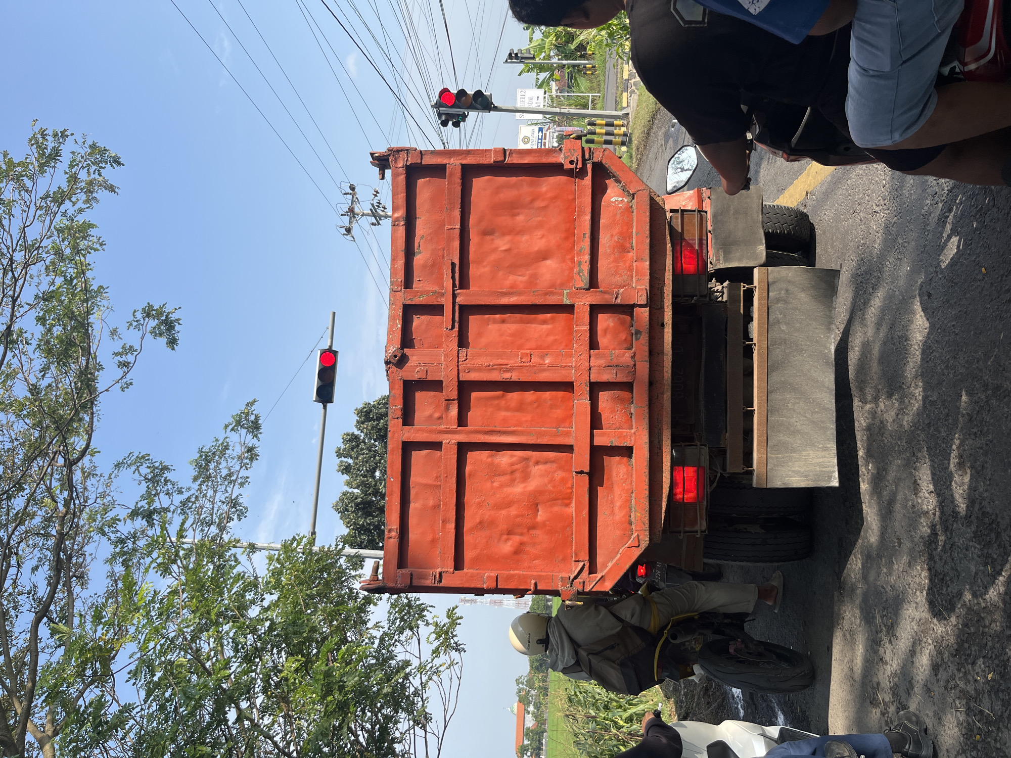 The image depicts a street scene in Getas Kidul, Indonesia, under a clear blue sky. The foreground is dominated by the rear of a large, orange dump truck. The truck's bed is visibly worn, showing signs of heavy use with a rough, unpainted texture. To the left of the truck, a person on a motorcycle, wearing a helmet and a backpack, is stopped. The motorcycle has a visible license plate. Further to the left, another person is partially visible, also on a motorcycle. To the right of the dump truck, another motorcycle is seen with two individuals. One of them, wearing a dark t-shirt and shorts, is looking forward. The other, wearing a blue shirt and light blue shorts, is seated behind. The road appears to be asphalt, with some debris and shadows cast by the vehicles and trees. In the background, a traffic light is visible and illuminated with a red signal. Overhead power lines and poles are present, along with trees and some distant buildings. A green field and a tower are also discernible in the far background. The scene suggests a traffic situation where vehicles are stopped, likely due to the red traffic light.