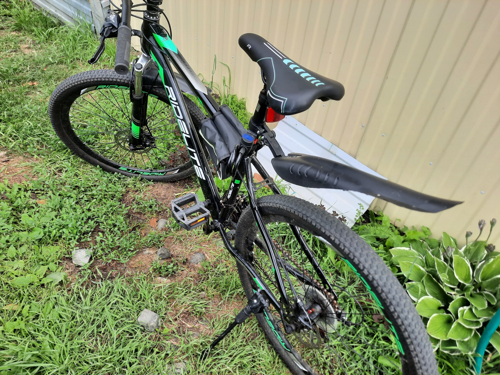 The image shows a black RIDE LITE bicycle parked on a grassy area next to a corrugated metal fence. The bicycle has green accents on the frame and wheels, a black saddle with teal accents, and a black kickstand. A black saddlebag is attached to the frame under the seat. The foreground is dominated by lush green grass and some rocks, while the background consists of the beige corrugated metal fence and some green foliage. The scene appears to be outdoors, likely in a residential area, given the presence of the fence and the cultivated plants. The lighting suggests it is daytime. There are no people or animals visible in the image. The location is specified as Bol'shoye Sorokino, Russia.