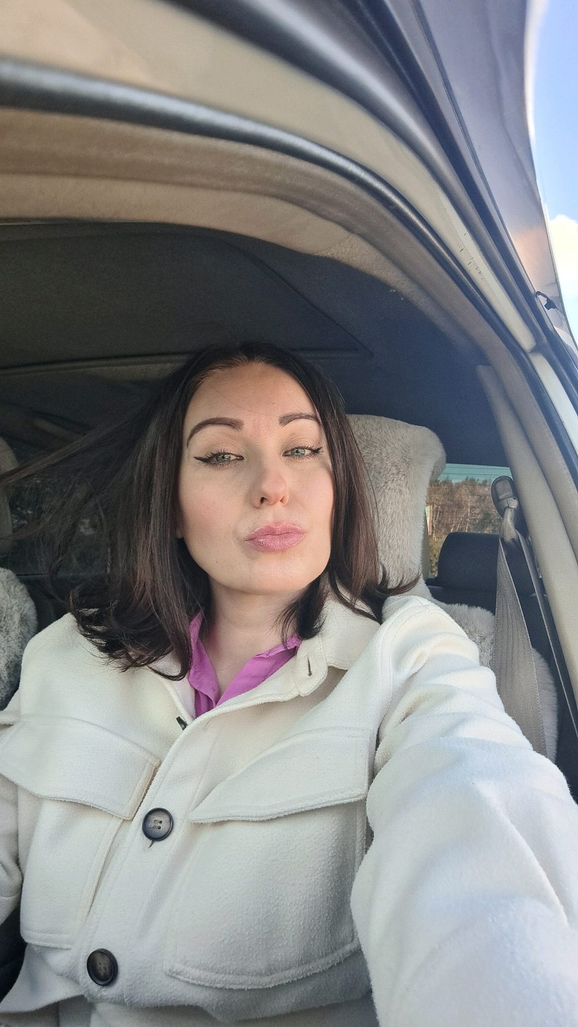 A woman with dark hair, green eyes, and heavy eyeliner is taking a selfie inside a car. She is wearing a white jacket over a pink shirt. Her lips are pursed. The car's interior is visible, including a fuzzy seat cover and a seatbelt. The sky is visible through the window, with some trees in the background. There is no information available to suggest the location is in Irkutsk, Russia.