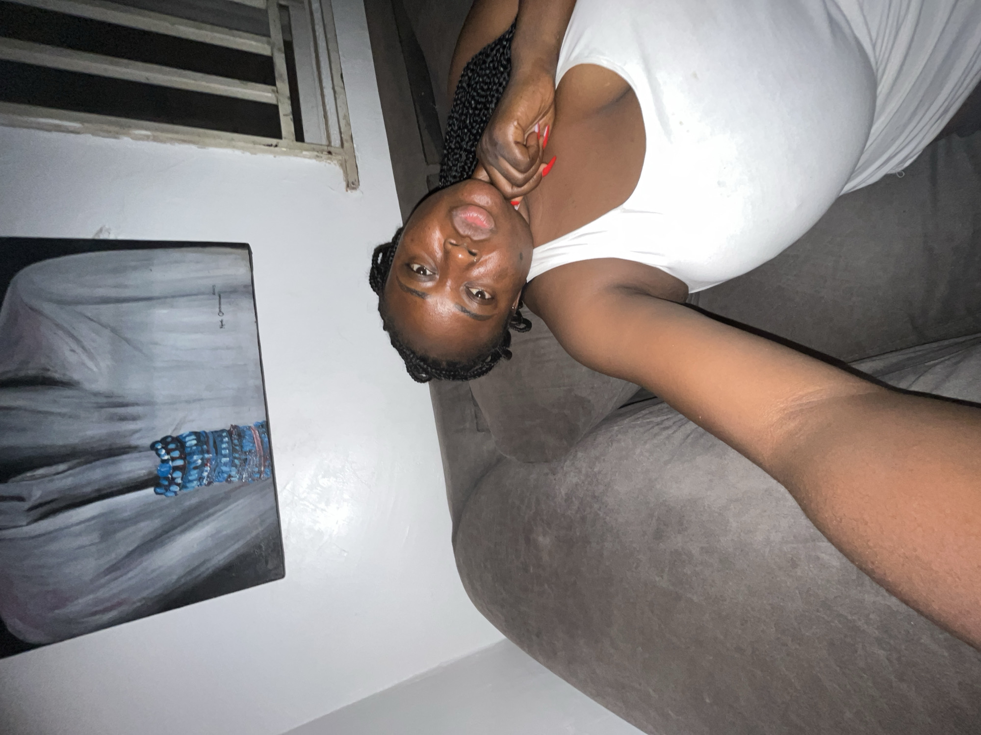 A woman lies on a gray couch. She wears a white shirt, has dark skin, and braided hair. Her right hand is raised to her neck, displaying red nails. Behind her to the left is a painting of a figure, primarily in shades of gray with a blue detail. Above the painting is a partially visible white wooden window frame. The setting appears to be indoors, likely a residence. While the location is speculated to be Abuja, Nigeria, there's no visual confirmation of the city in the image.