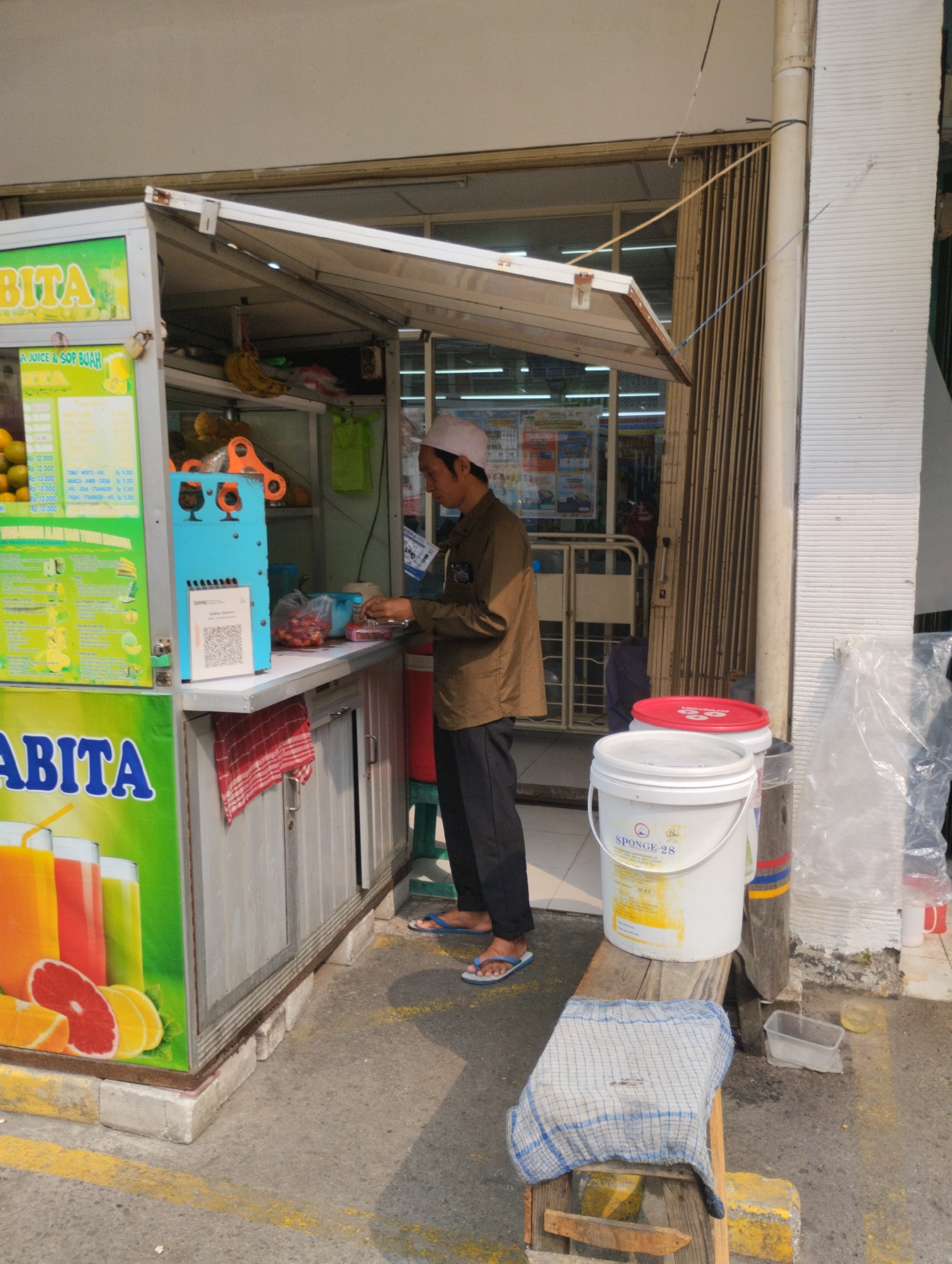 A man wearing a white peci and a brown long-sleeved shirt is seen preparing drinks at an outdoor juice stall. The stall has a blue juicing machine and various fruits displayed. A sign on the stall reads "BITA JUICE & SOP BUAH". In the background, a folded metal gate and the interior of a store are visible. Two white buckets, one labeled "SPONGE-28", are placed next to the stall. The scene appears to be in an urban environment, possibly a marketplace or sidewalk, in the city of Bekasi, Indonesia. The ground is paved, with yellow lines indicating a pedestrian or traffic boundary.
