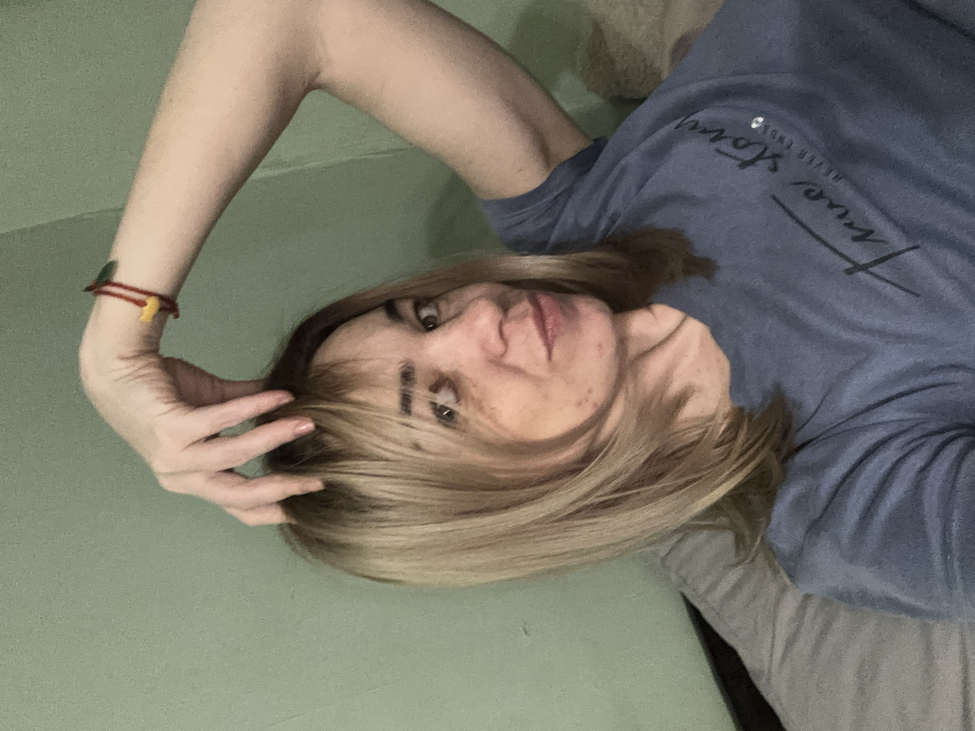 A woman with blonde hair and a blue T-shirt is lying down. She is touching her hair with her right hand. A red bracelet is visible on her wrist. The background is a green wall and a grey surface. The image doesn't contain enough information to determine the location is in Vsevolozhsk, Russia.