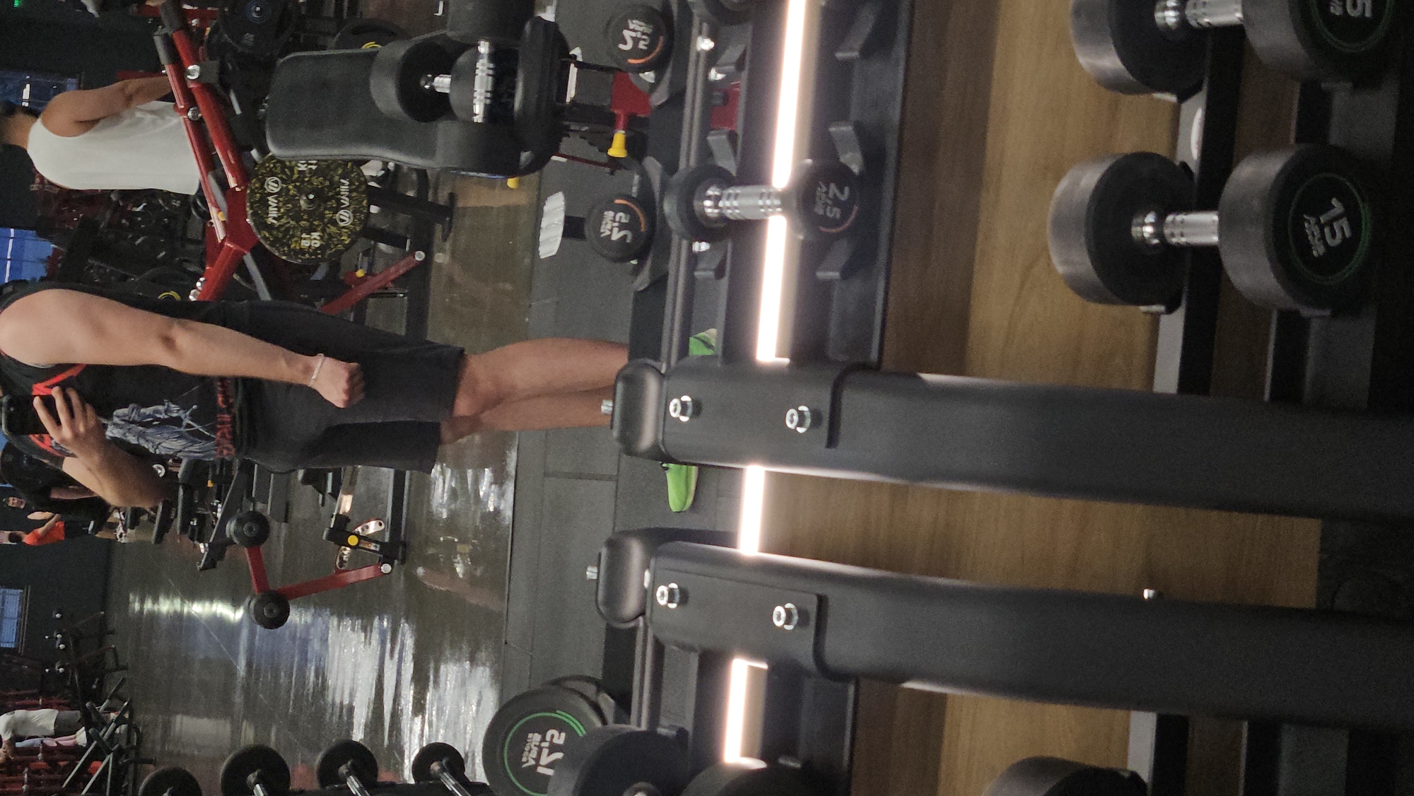 The image depicts an indoor gym located in Plovdiv, Bulgaria. The primary subject is a man taking a mirror selfie. He is positioned in the left foreground (when oriented correctly), wearing a black short-sleeved t-shirt with an indistinct graphic design and black shorts, holding a phone to capture his reflection. His reflection is visible in the reflective surface that occupies the left side of the frame.
In the background, another man is partially visible, wearing a white tank top and seemingly engaged with a red piece of gym equipment. The gym floor is dark and shows reflections from the overhead lighting.
Various gym equipment is present. On the right side of the image, a robust black dumbbell rack is prominently displayed, featuring integrated vertical LED light strips that illuminate the rack and the wooden wall behind it. Several dumbbells are neatly arranged on the rack. In the middle ground and background, various weight plates are visible; some are gold and black, marked with "20 KG" and "VARNA", while a green and black plate in the lower left corner shows "K2".
Visible text on the dumbbells includes "15" and "2.5", indicating their respective weights. The lighting is artificial, characteristic of an indoor fitness facility, creating bright reflections, particularly on the floor and from the dumbbell rack's light strips. The time of day is not discernible due to the indoor setting.