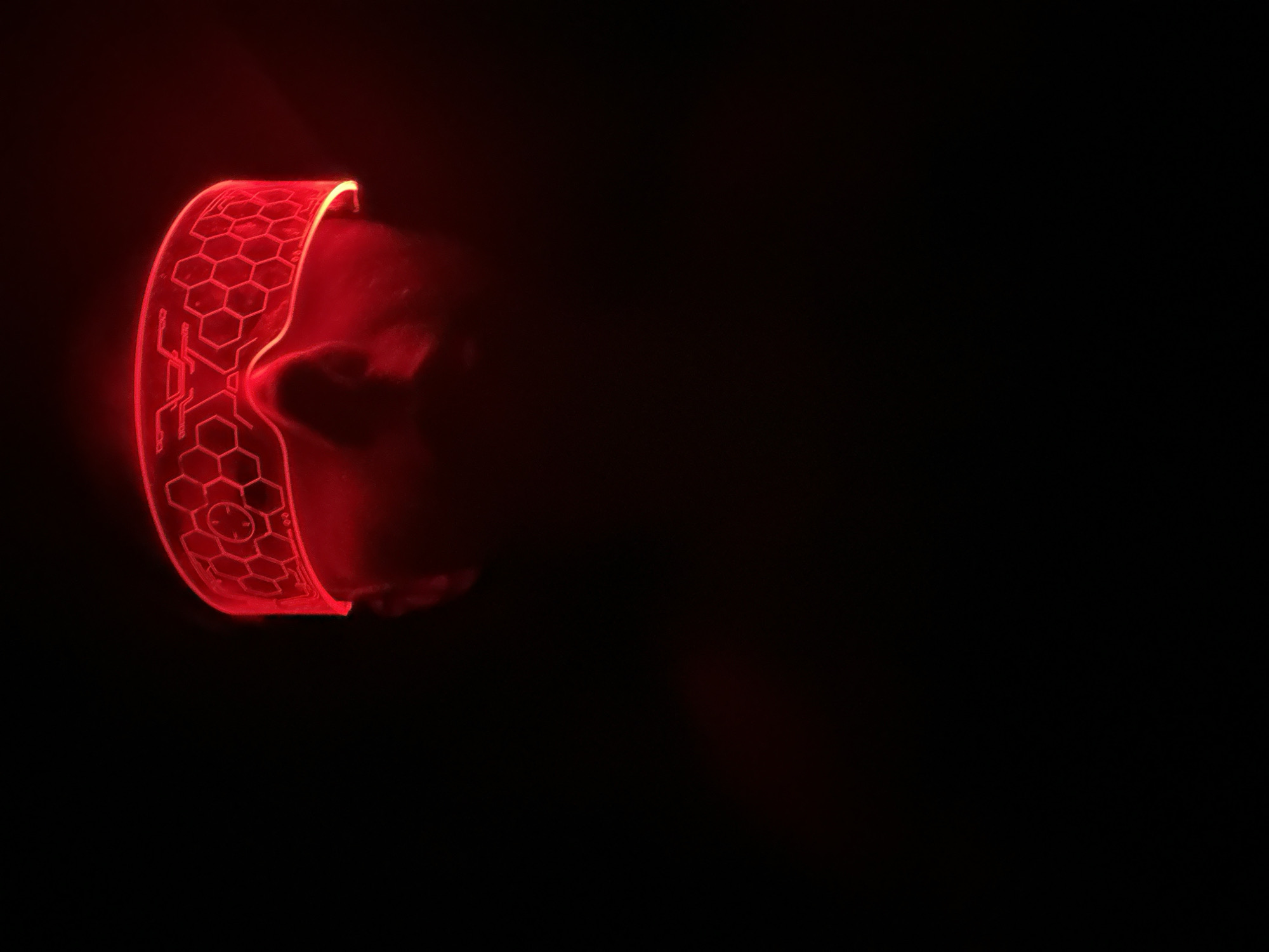 The image displays a close-up of a person's head in a profoundly dark environment, likely an indoor setting at night in Votkinsk, Russia. The central subject is a glowing red headband or visor worn across the individual's forehead and eyes. This translucent object emits a vibrant red light, which illuminates its intricate patterns and casts a soft, deep red glow onto the wearer's face.
The visible part of the person's face, a partial profile, is largely obscured by shadow and the intense glow, revealing only faint outlines of an eye, nose, and cheek. The headband features a striking design composed of interconnected hexagonal shapes resembling a honeycomb, interspersed with circuit-like lines and geometric elements, contributing to a futuristic or high-tech aesthetic. No specific activity beyond wearing the device is depicted. The surrounding space is entirely black, drawing full attention to the luminous red object and creating a mysterious and enigmatic atmosphere. No visible text can be discerned on the object or in the background.