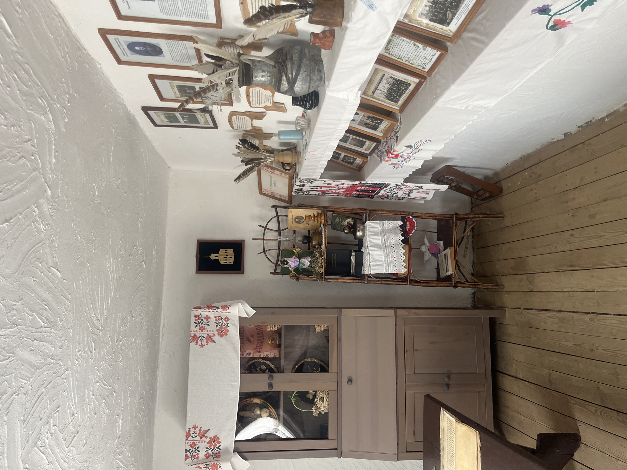 The image depicts an interior scene, likely within the Taman Museum in Taman', Russia, exhibiting a collection of artifacts and decor that evoke a historical or ethnographic theme. The room features a textured white ceiling and white walls. On the left, a tall, two-section wooden cabinet with glass doors on the upper section displays various decorative plates and baskets. A tablecloth with a red floral embroidered pattern is draped over the top of the cabinet. To the right of the cabinet is a narrow, multi-tiered shelving unit made of dark wood, adorned with books, a doll, dried plants, a kerosene lamp, and a decorative item resembling a small church.
The main wall on the right is lined with a long shelf covered by a white tablecloth, also featuring embroidered floral patterns. This shelf holds an assortment of items, including a large metal vase filled with feathers, smaller vases, candles, a small black ceramic object, and a red decorative piece. Mounted on the wall above the shelf are numerous framed pictures and documents, many with wooden plaques. A prominent display of framed historical photographs and documents is arranged on the shelf itself, covered in part by a red and white embroidered fabric.
The floor is made of light-colored wooden planks. In the foreground on the left, a dark wooden table with a document or book on its surface is partially visible. The overall impression is a meticulously arranged display of historical artifacts and decorative items, suggesting a preserved interior or exhibit.