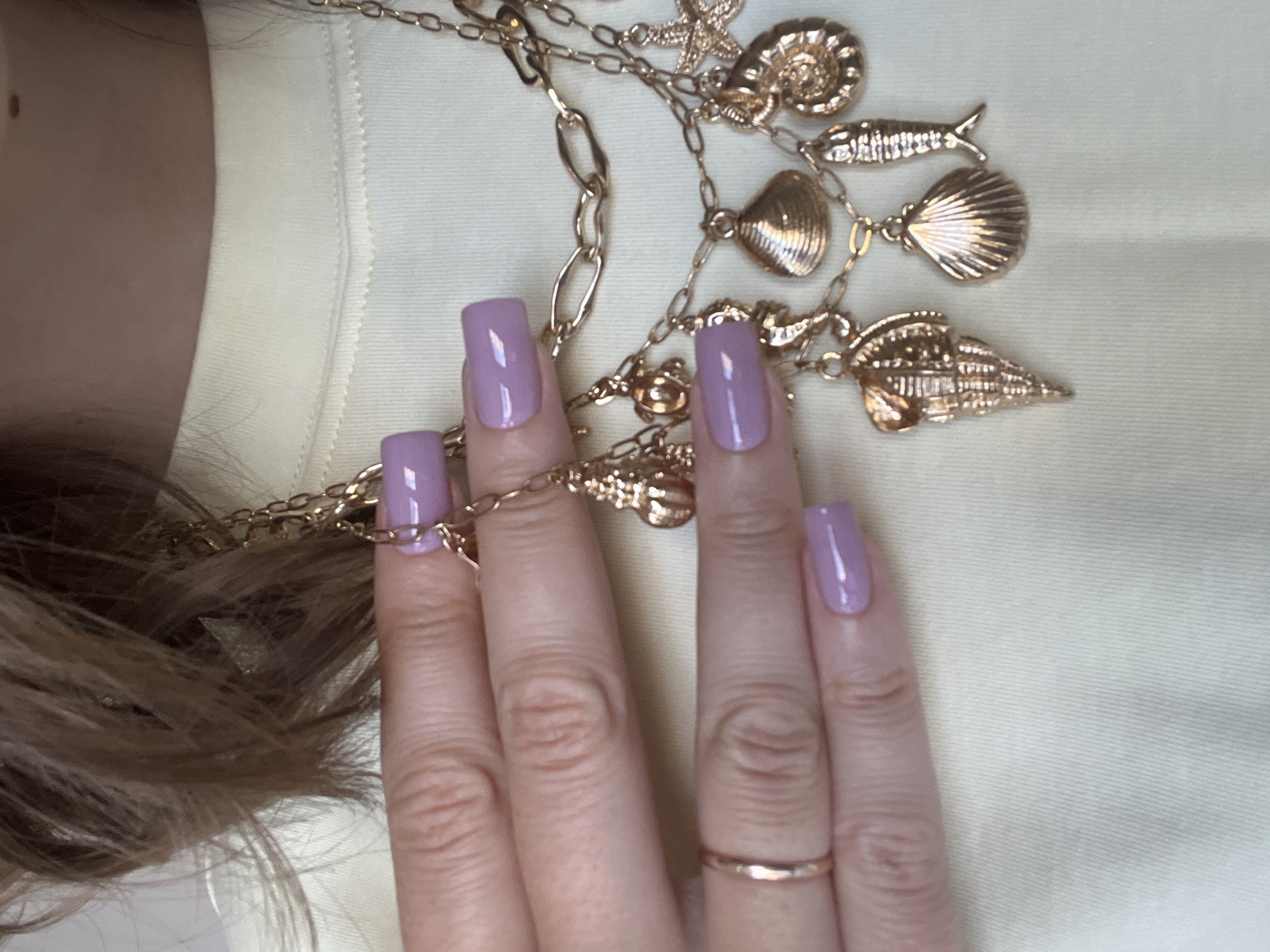 A close-up shot shows a person's hand with lavender-colored square-shaped fingernails. The hand is adorned with a thin, gold ring on one finger and rests against a light yellow or off-white background. A gold chain necklace with various sea-themed charms such as starfish, shells, and a fish, is draped across the hand and the background. Part of the person's blonde hair is visible on the left side of the frame. The location of the image is not discernible as Podgorica, Montenegro, or any other specific location.