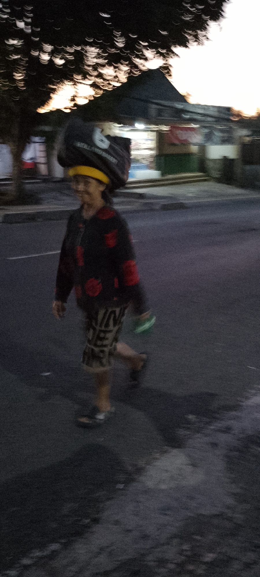 In Pekutatan, Indonesia, an adult is captured in motion, walking along a paved road under a twilight sky, the foliage of a nearby tree blurred with streaks of light. The individual, seen from the waist down, is wearing a black and red patterned long-sleeved shirt over patterned shorts. Their right hand appears to be holding a small, green object. A large, dark bag with a yellow band rests precariously on their head. The background suggests an urban or semi-urban setting with the faint outlines of buildings and streetlights. The lighting indicates either early morning or late evening, with a warm glow emanating from above and behind.