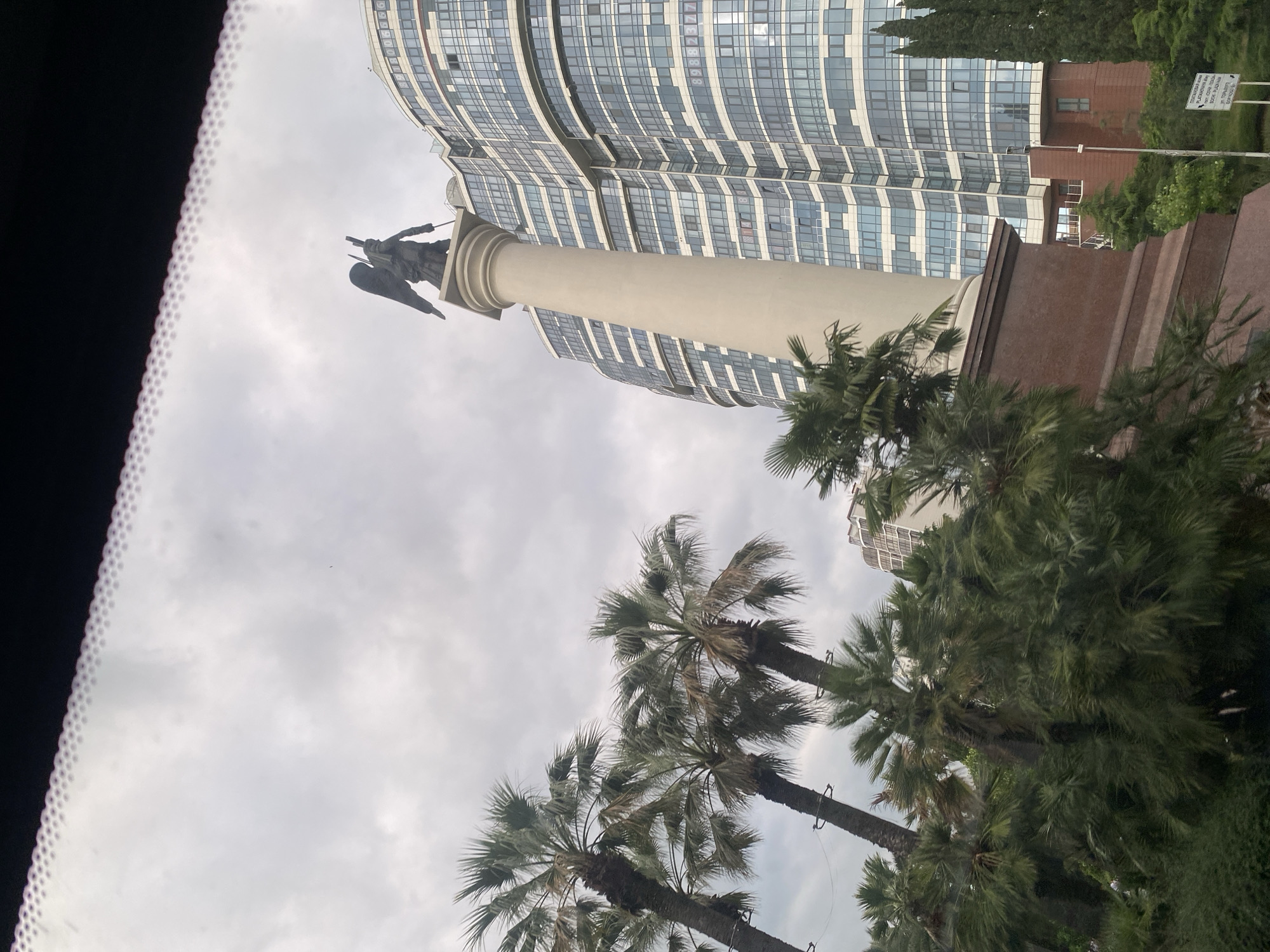 The image depicts a cloudy day in Sochi, Russia, with a view of a modern glass-facade building and a monument. The building, characterized by its numerous windows and curved design, dominates the background. In the foreground, a tall, light-colored column supports a dark, winged statue, possibly an angel or a winged victory, holding a spear. Palm trees, with their characteristic fronds, are scattered throughout the scene, suggesting a warm climate. The sky is overcast, casting a diffused light over the area.
To the right of the monument and building, a white sign with black text is visible, though the text is mostly illegible due to distance and image resolution. The overall impression is one of an urban environment with historical or symbolic elements, set against a backdrop of lush vegetation and a somewhat somber sky. The composition, with the camera angled upwards, emphasizes the scale of the building and the monument. The presence of palm trees and the modern architecture hint at a coastal or resort-like setting, consistent with Sochi's reputation. There is no discernible activity or event taking place; the image appears to be a still shot of the location.