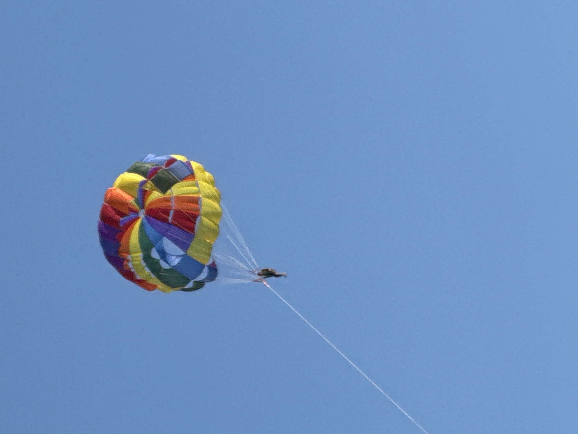 A single person is parasailing against a clear blue sky. The parasail is multicolored, with panels of red, orange, yellow, green, blue, and purple. A thin white line extends from the parasail down to the person, who is wearing dark clothing and appears to be in a seated position. The parasail is positioned towards the upper right of the frame, with the person and tow line extending diagonally down towards the lower left. The image appears to have been taken in Budva, Montenegro, a coastal city known for its beaches and water activities.