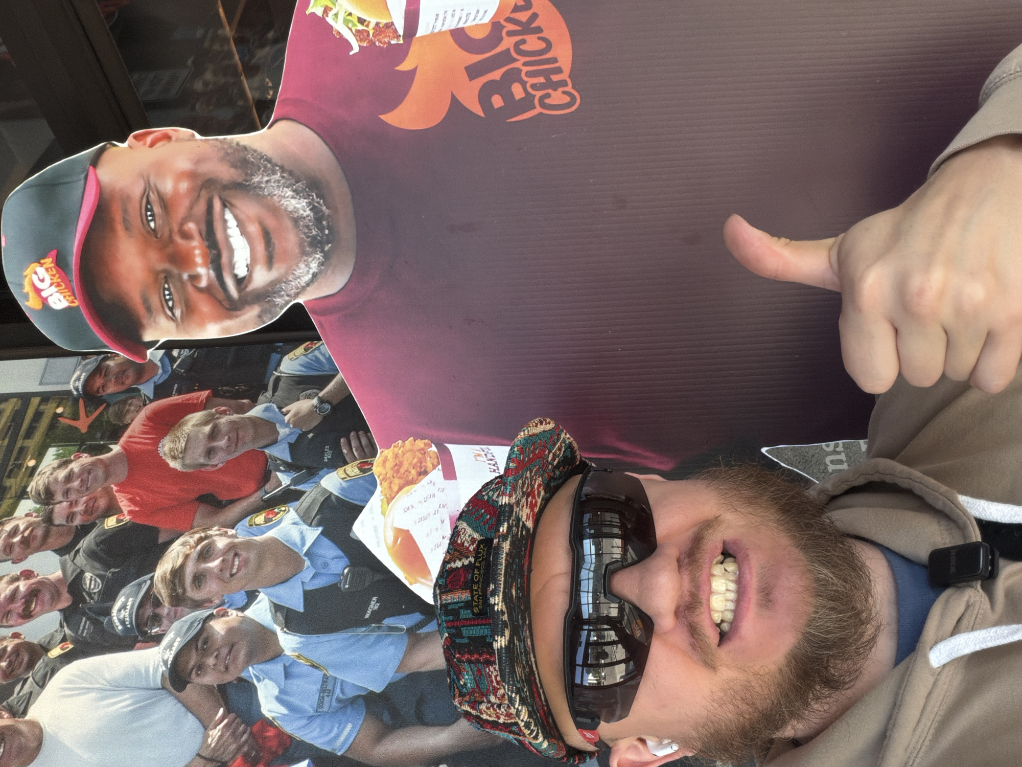 The image shows a man taking a selfie with a promotional display for "Big Chicken" in Rosemont, Illinois, USA. The man is wearing a patterned hat, sunglasses, and a light brown hoodie. He has a beard and is smiling broadly, displaying his teeth. He is giving a thumbs up with his right hand.

In the background are two cardboard cutouts, a cardboard cutout of Shaquille O’Neal dressed in a maroon shirt with a Big Chicken logo, and a cardboard cutout of an image featuring multiple police officers. A box of Big Chicken product is also visible. Text on the cardboard includes the words "STATE OF FLUX SAN FRANCISCO" and "BIG CHICKEN." A black device is clipped to the hoodie.