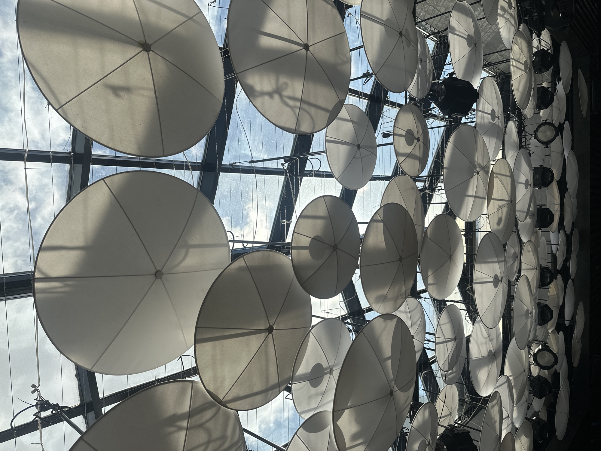 The image shows a ceiling installation of numerous white, circular parasols suspended in a grid-like pattern within a glass-roofed structure. The parasols are translucent, allowing light to filter through, and they are mounted on what appear to be metal frames. Some lighting fixtures are interspersed among the parasols, suggesting the structure serves as a lighting fixture itself. Through the glass roof, one can see a pale blue sky with some cloud cover. The overall perspective is from below, looking upward toward the ceiling. The location is most likely an interior space, judging from the artificial lighting and engineered design of the ceiling. Specific details to pinpoint the location in Izhevsk, Russia, are not apparent.