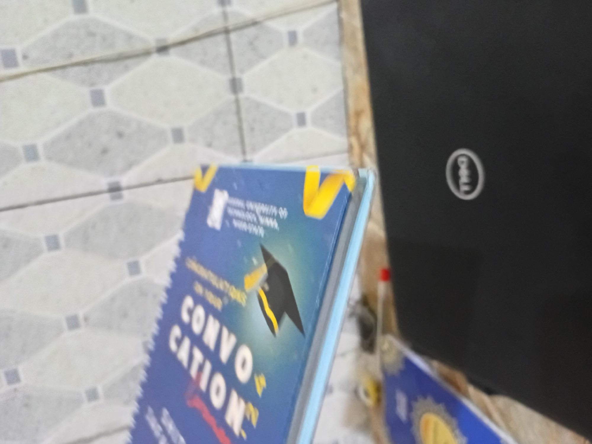 The image shows a partial view of what appears to be a book with the word "CONVOCATION" prominently displayed on its blue cover, alongside a black graduation cap graphic. Adjacent to the book is a black Dell laptop. The background includes a patterned tile surface. The location is Unknown, Unknown.