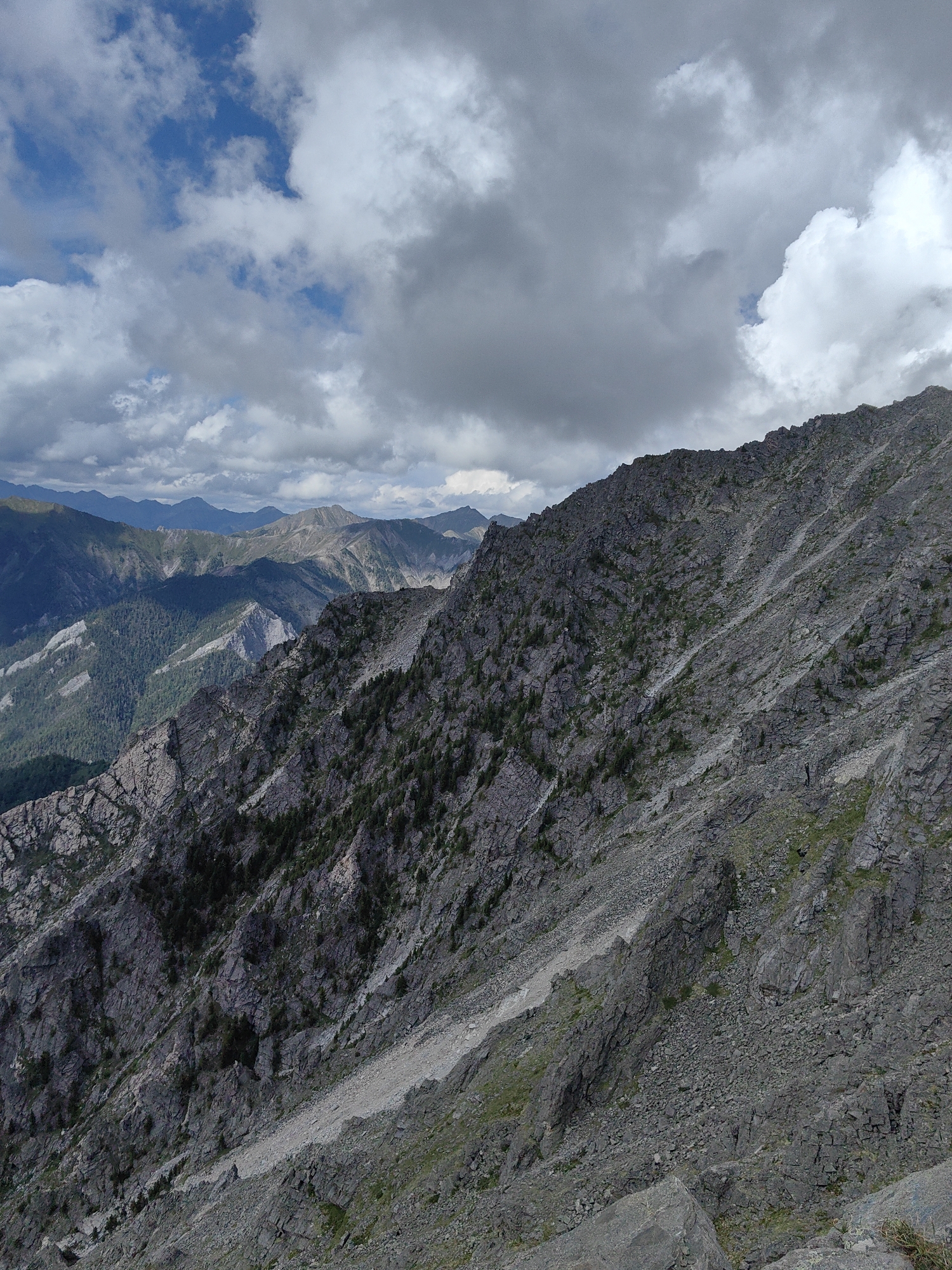 This is a high-angle, full outdoor shot of a mountainous landscape under a cloudy sky. The foreground is dominated by a steep, rocky slope covered in scree and sparse vegetation, with scattered dark green pine trees. The slope recedes into the distance, revealing layers of more distant, green, and grey mountains bathed in shadow and sunlight. The sky above is filled with a mix of dark, heavy clouds and brighter, puffy white clouds, with patches of blue sky visible. There are no people or discernible structures in the image. This scene is consistent with the natural, undeveloped mountainous terrain found in the vicinity of Arshan, Russia, known for its alpine scenery.